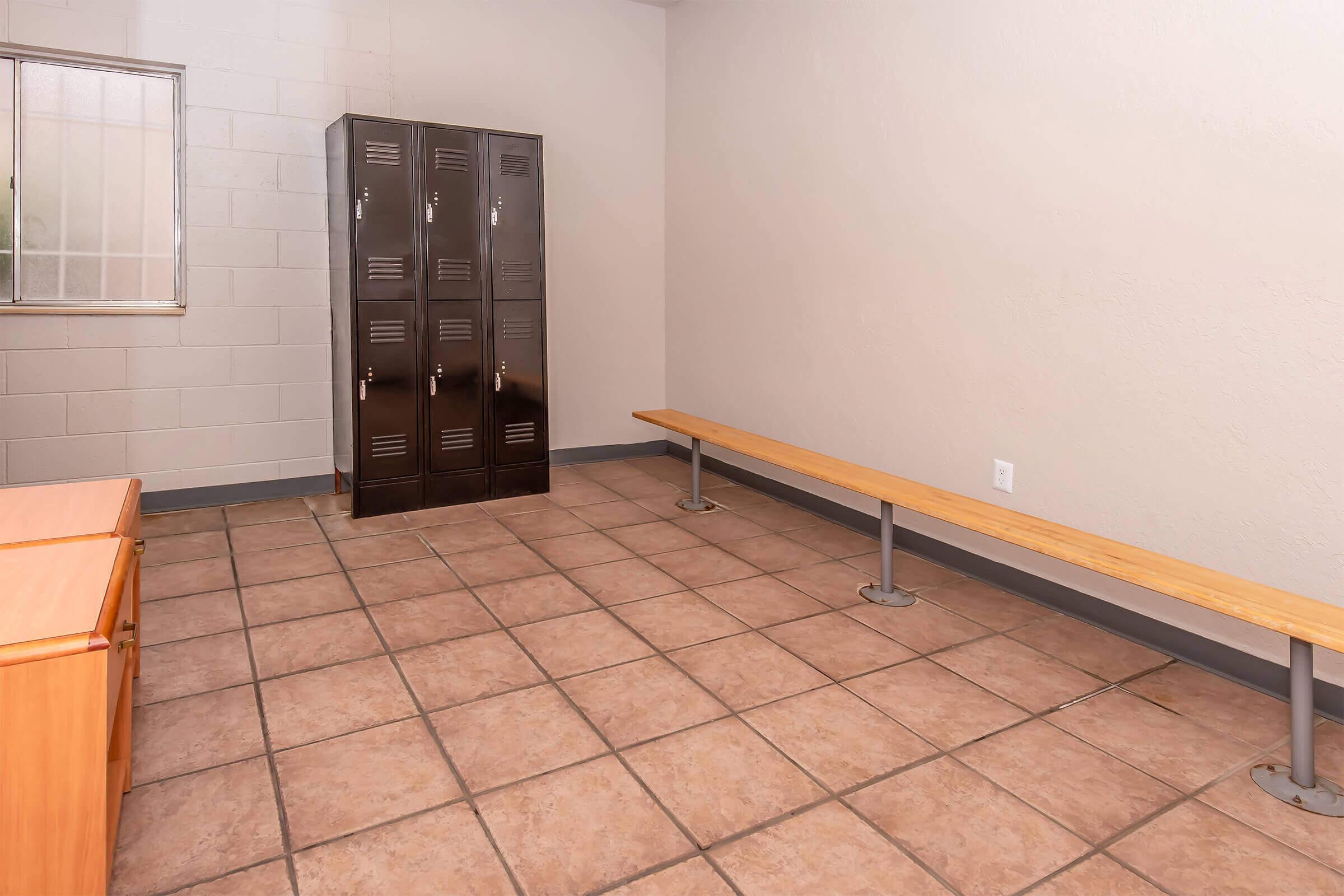 A sparsely furnished room with beige walls and tiled flooring. On the left, there is a wooden bench, while to the right, a set of black lockers stands against the wall. A window lets in natural light, illuminating the space. The overall atmosphere is utilitarian and simple.