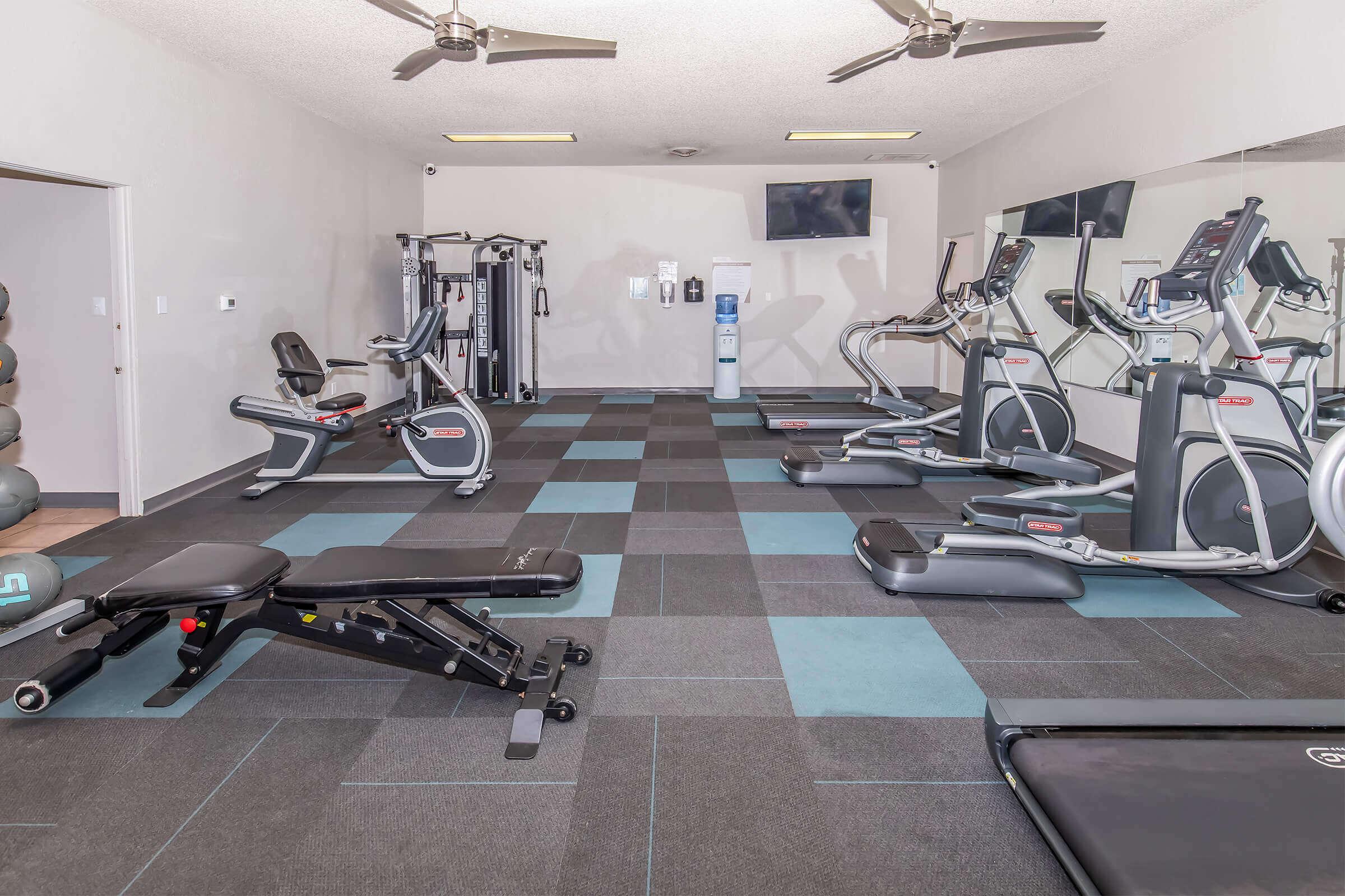 A well-equipped gym featuring various exercise machines, including treadmills, stationary bikes, and a weight bench. The floor is covered with black and blue tiles, and there are ceiling fans and two wall-mounted TVs. The space is well-lit and organized for fitness activities.