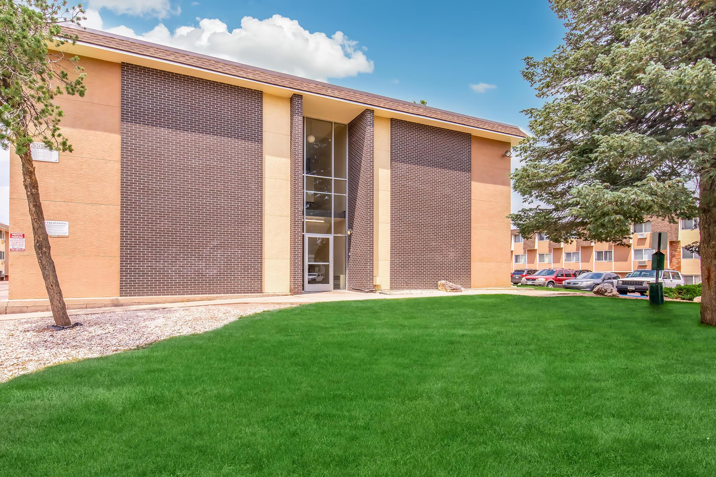 A modern multi-story building with a brown brick facade and large glass entrance, surrounded by a well-maintained green lawn. Pine trees are visible on the side, and additional buildings can be seen in the background. The sky is clear with a few fluffy clouds.