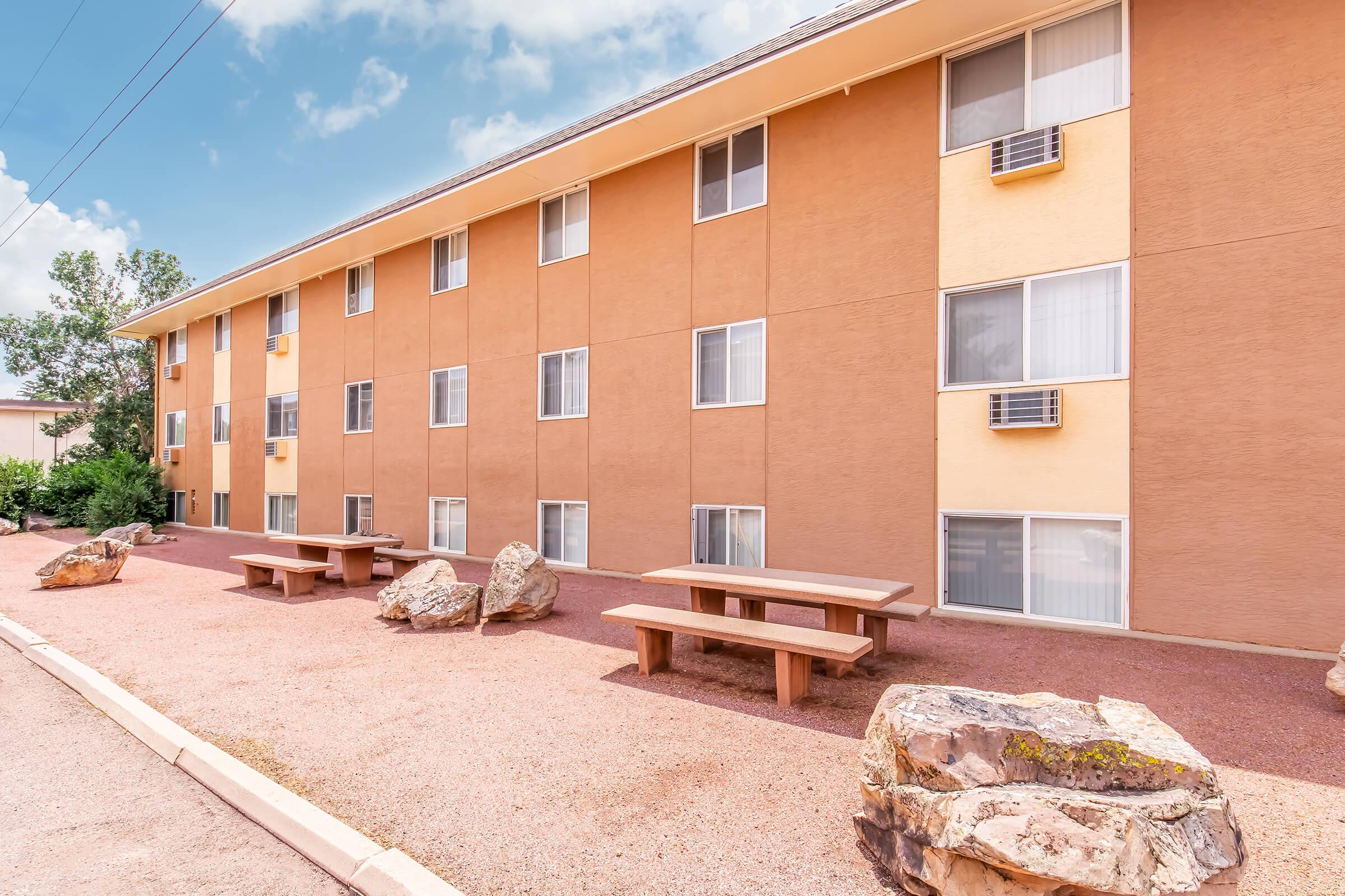 A view of a multi-story apartment building in a tan color, featuring several windows and air conditioning units. In the foreground, there are picnic tables and large rocks on a gravel surface, surrounded by greenery and blue sky.