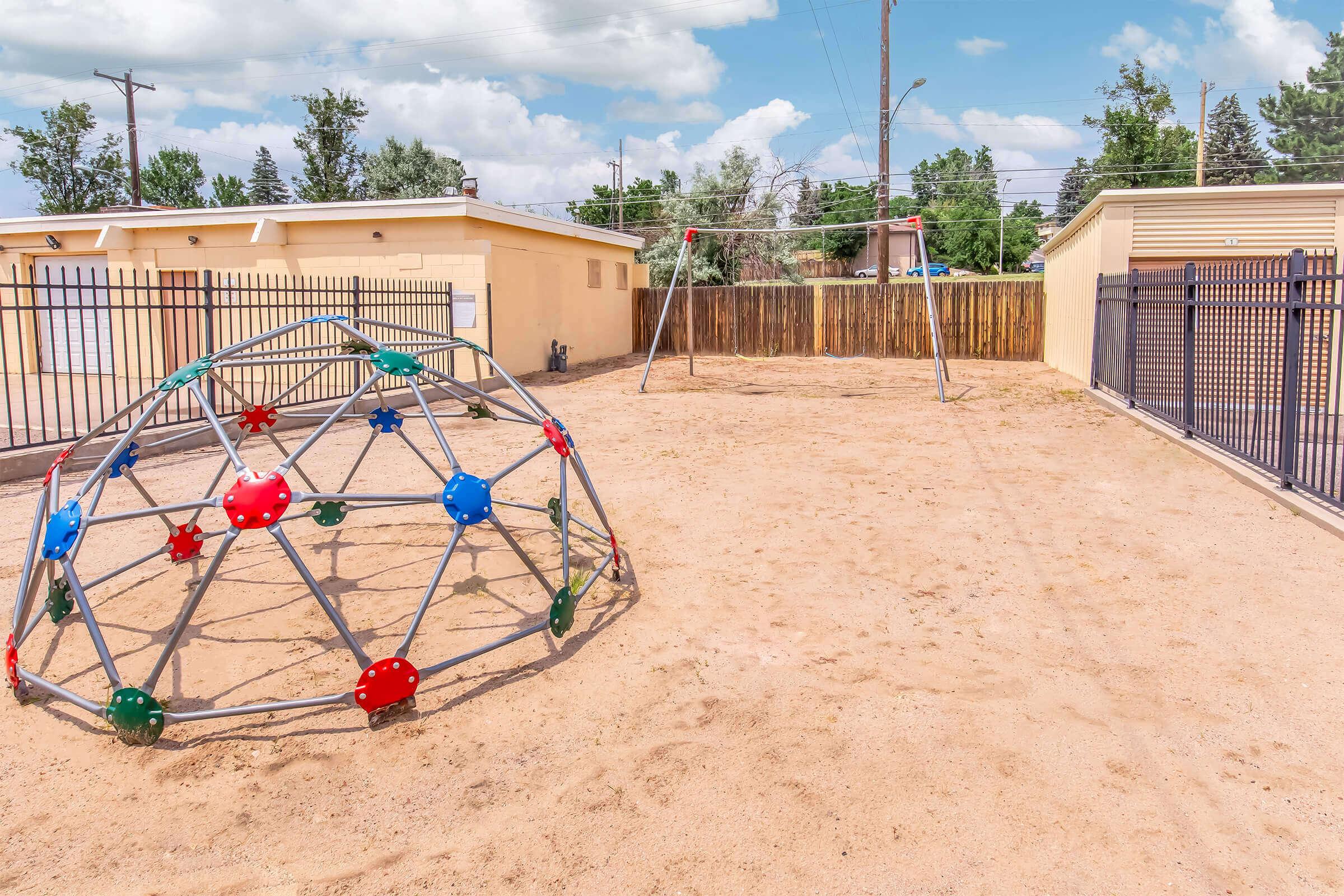 A playground area featuring a colorful climbing dome and two swings set against a backdrop of beige buildings and a clear blue sky. The ground is sandy, and there are trees in the distance, creating a bright and inviting outdoor space for children to play.