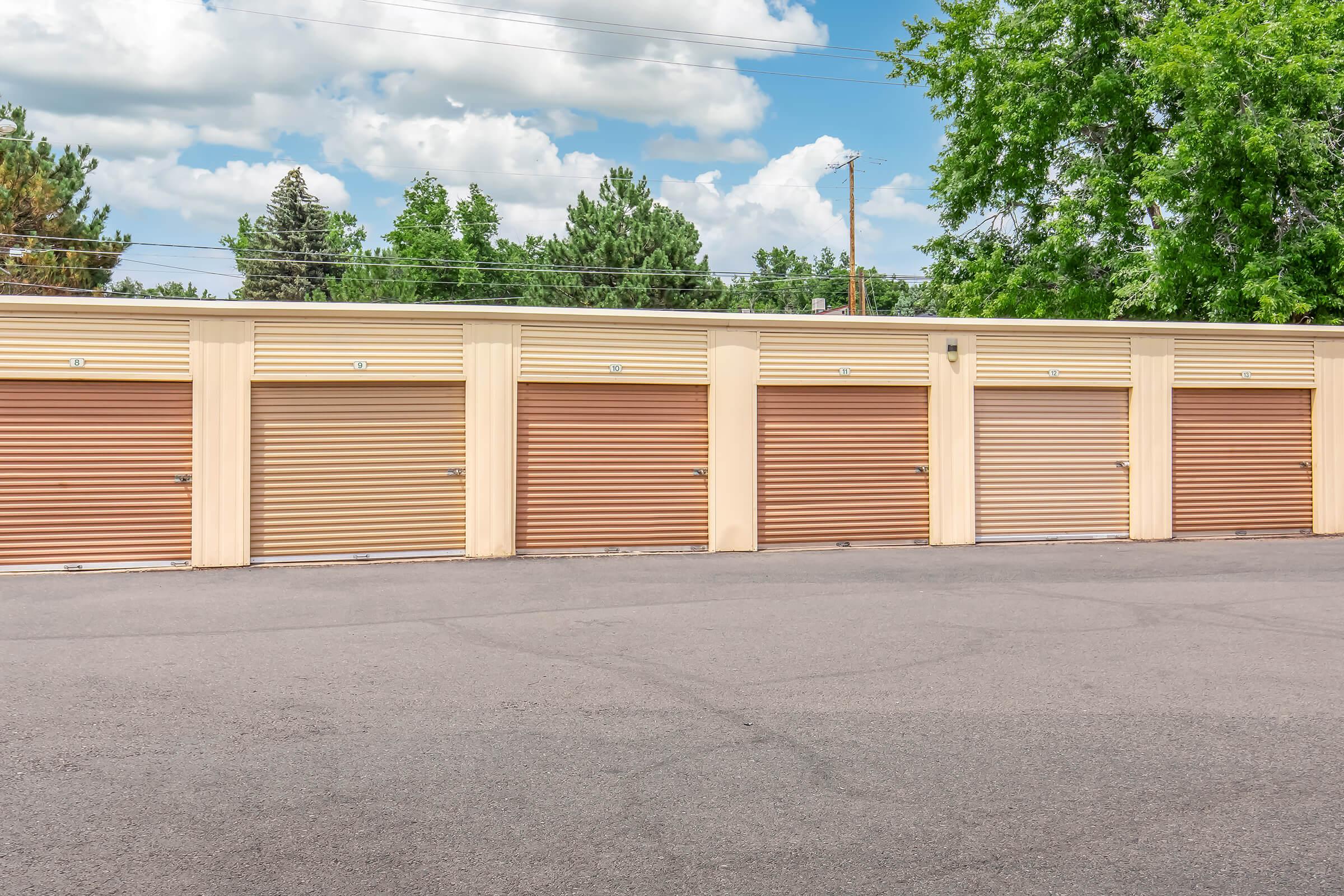 A row of storage units with tan and brown doors, set against a backdrop of green trees and a partly cloudy sky. The asphalt ground in front of the units is smooth and well-maintained. There are several storage doors visible, each with a different color, indicating individual units.