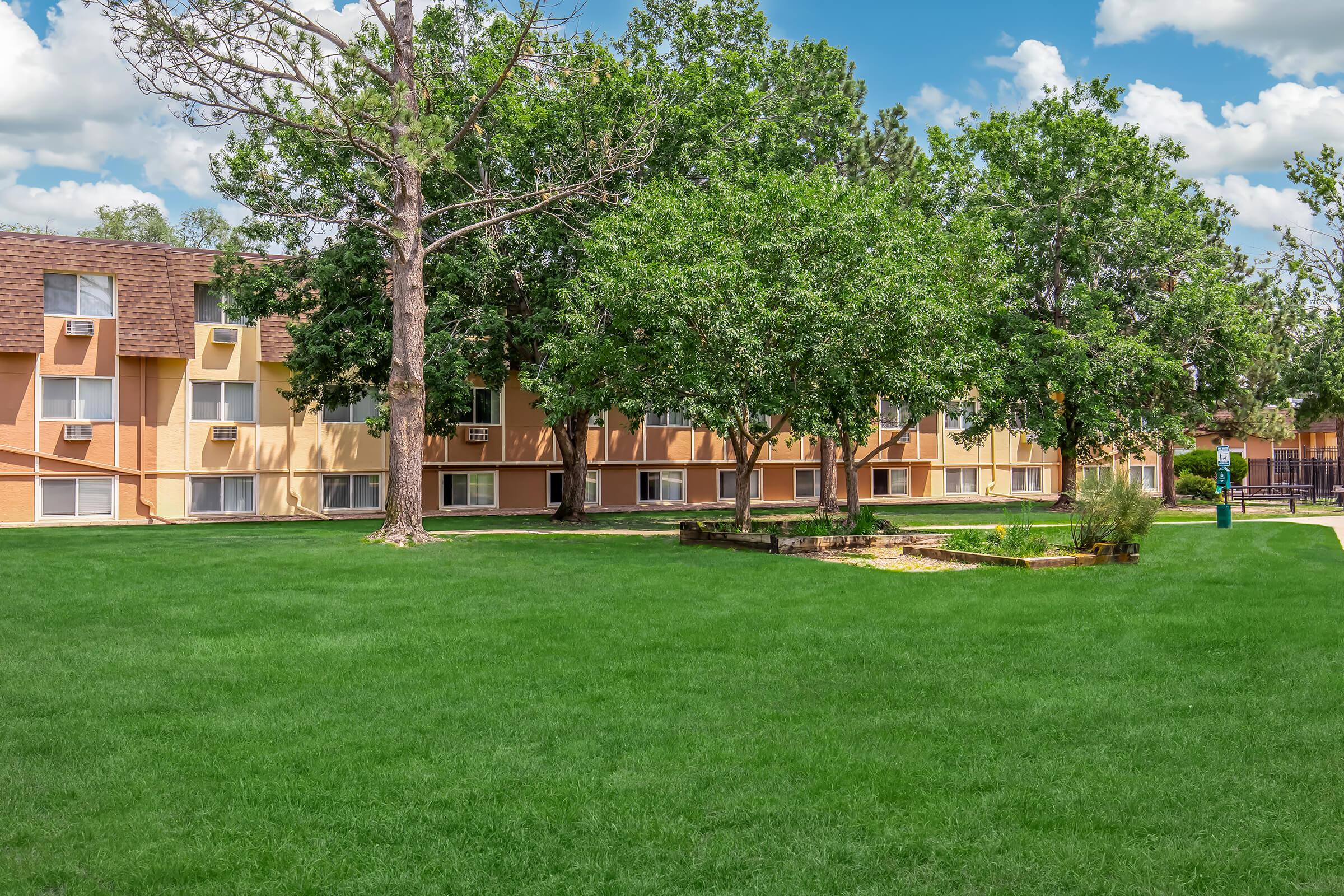 A well-maintained residential building with multiple levels, surrounded by lush green grass and large trees. The scene captures a sunny day with a blue sky and fluffy clouds. A small landscaped area with stones and shrubs can be seen in front of the building, adding to the outdoor aesthetic.