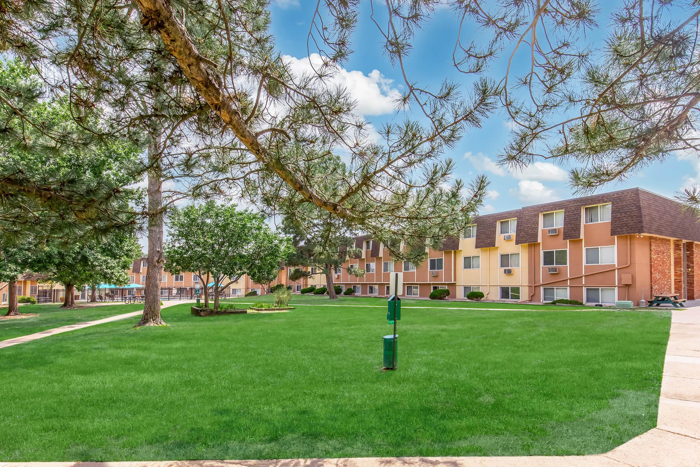 A well-maintained residential area featuring a green lawn surrounded by trees and shrubs. In the background, there are two-story apartment buildings with balconies. The sky is clear with some clouds, creating a bright and inviting atmosphere.