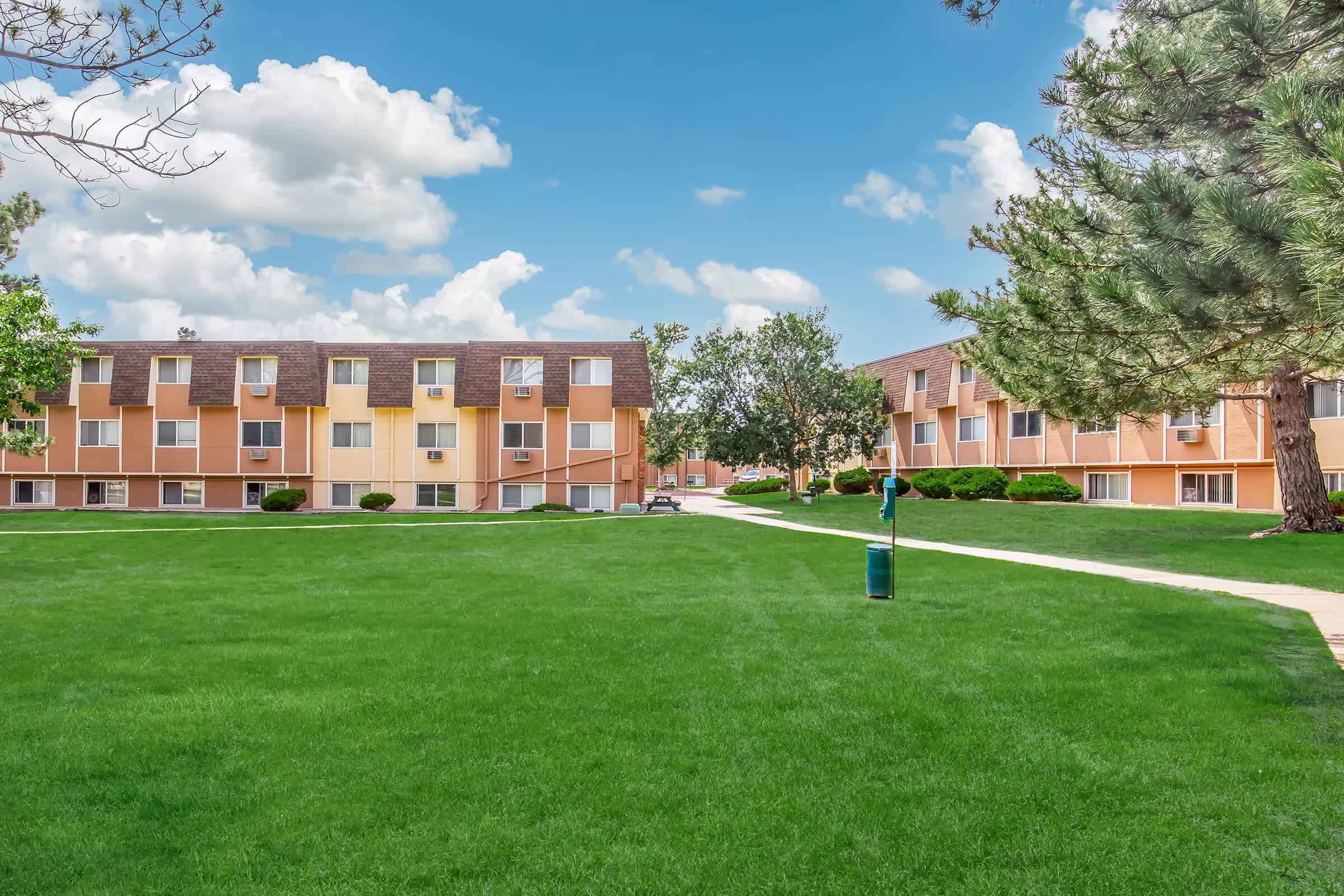 A view of a well-maintained apartment complex featuring two buildings with orange and beige exteriors. The grassy area is lush and green, with a few trees and a path leading through the lawn. The sky is bright blue with fluffy white clouds, creating a welcoming environment.