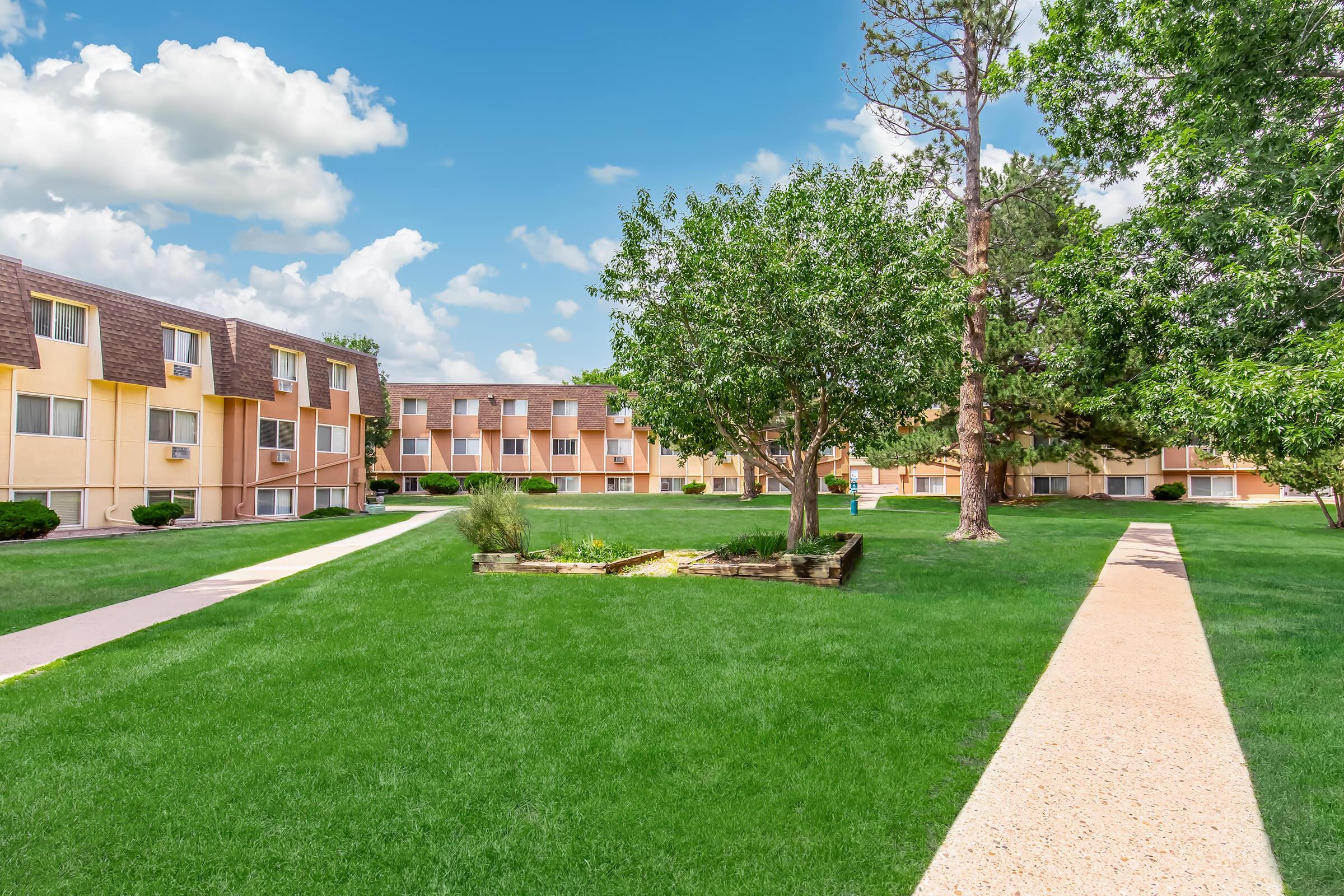 A well-maintained residential complex featuring two-story buildings surrounded by lush green grass, trees, and landscaped walkways. The sky is bright with scattered clouds, creating a cheerful atmosphere. Pathways lead through the open area, enhancing the inviting and peaceful environment.