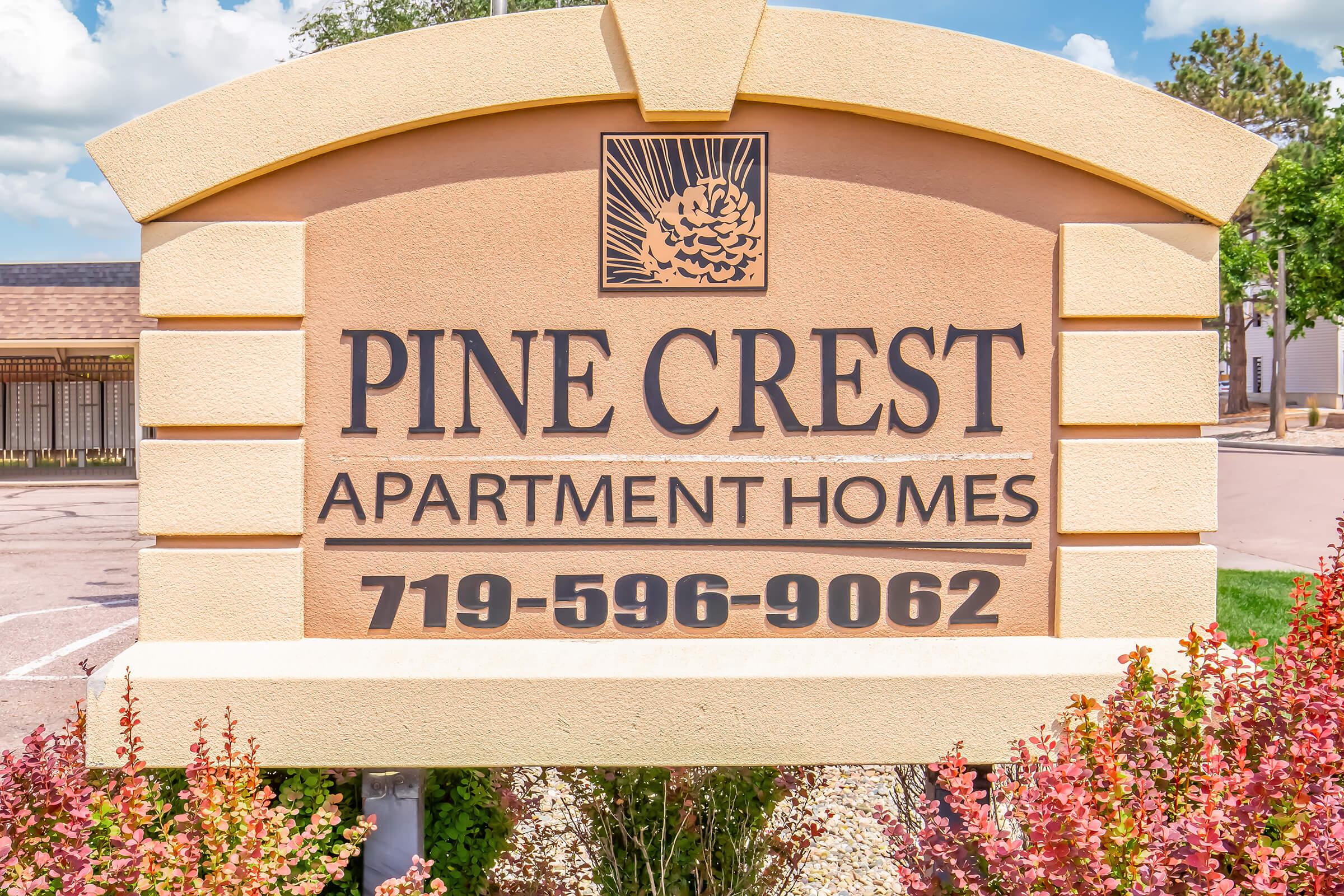Sign for Pine Crest Apartment Homes, featuring the name "Pine Crest" prominently, along with the text "Apartment Homes" and a phone number: 719-596-9062. The sign has a decorative design and is surrounded by colorful landscaping under a bright blue sky.