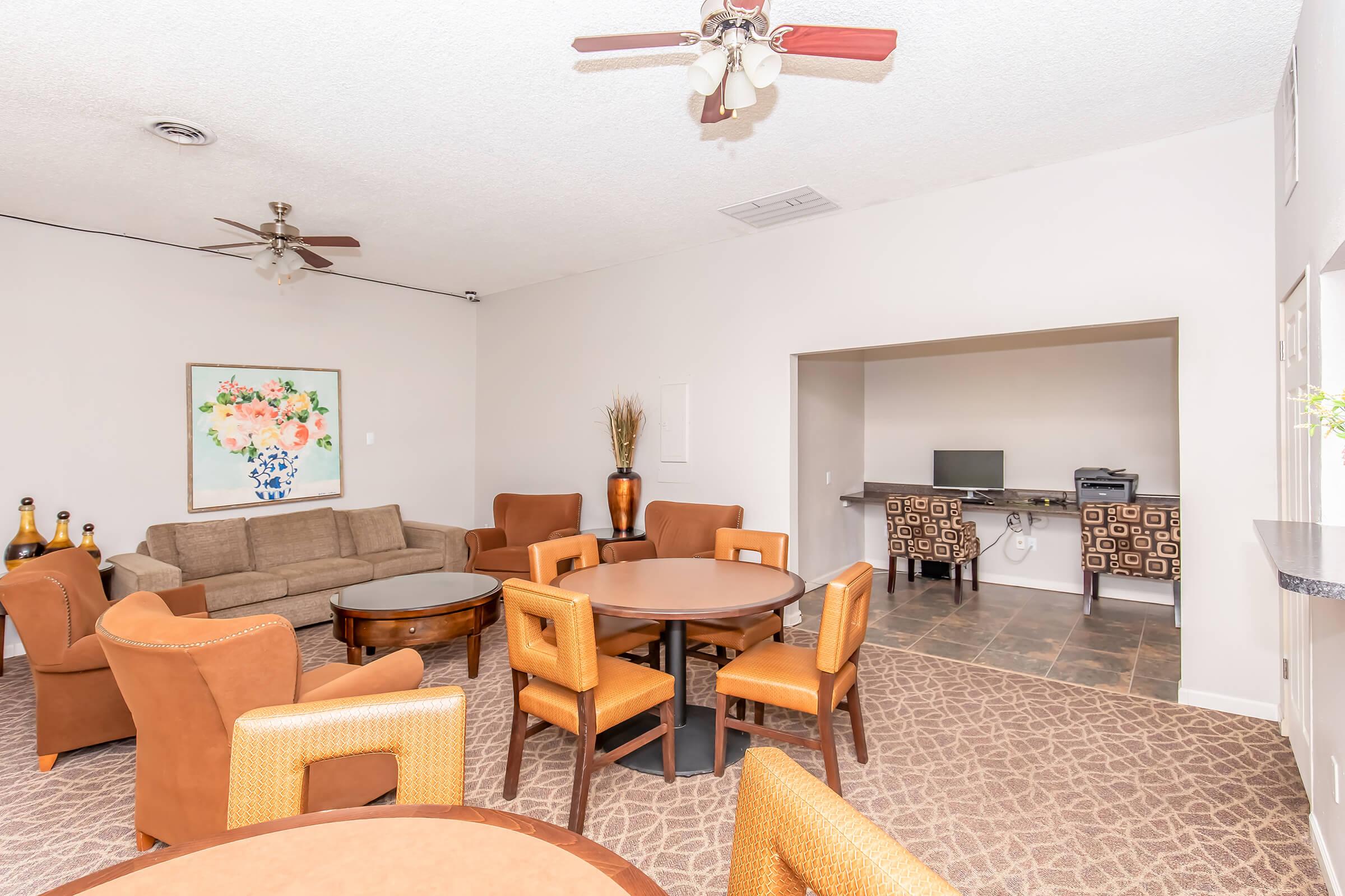 Spacious common area featuring a cozy seating arrangement with a beige couch and circular wooden tables surrounded by brown chairs. A ceiling fan is visible, and a large floral painting decorates one wall. In the background, there is a kitchenette or bar area with additional seating and a television.