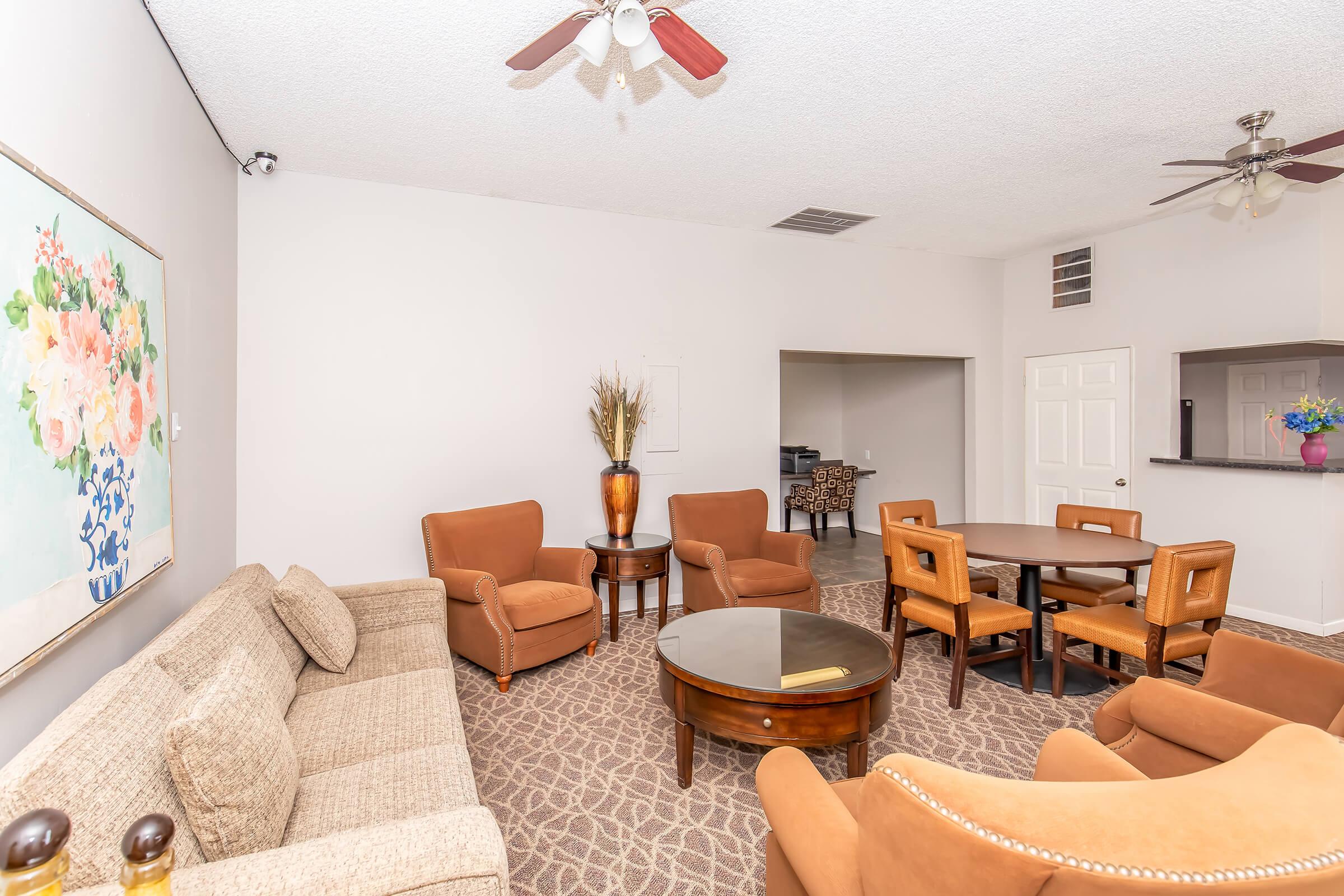 A spacious living area featuring a beige couch, two brown armchairs, and a round wooden coffee table. There's a dining table with four chairs in the background. A floral painting is displayed on the wall, and a ceiling fan is visible. The decor is light and inviting, creating a cozy atmosphere.
