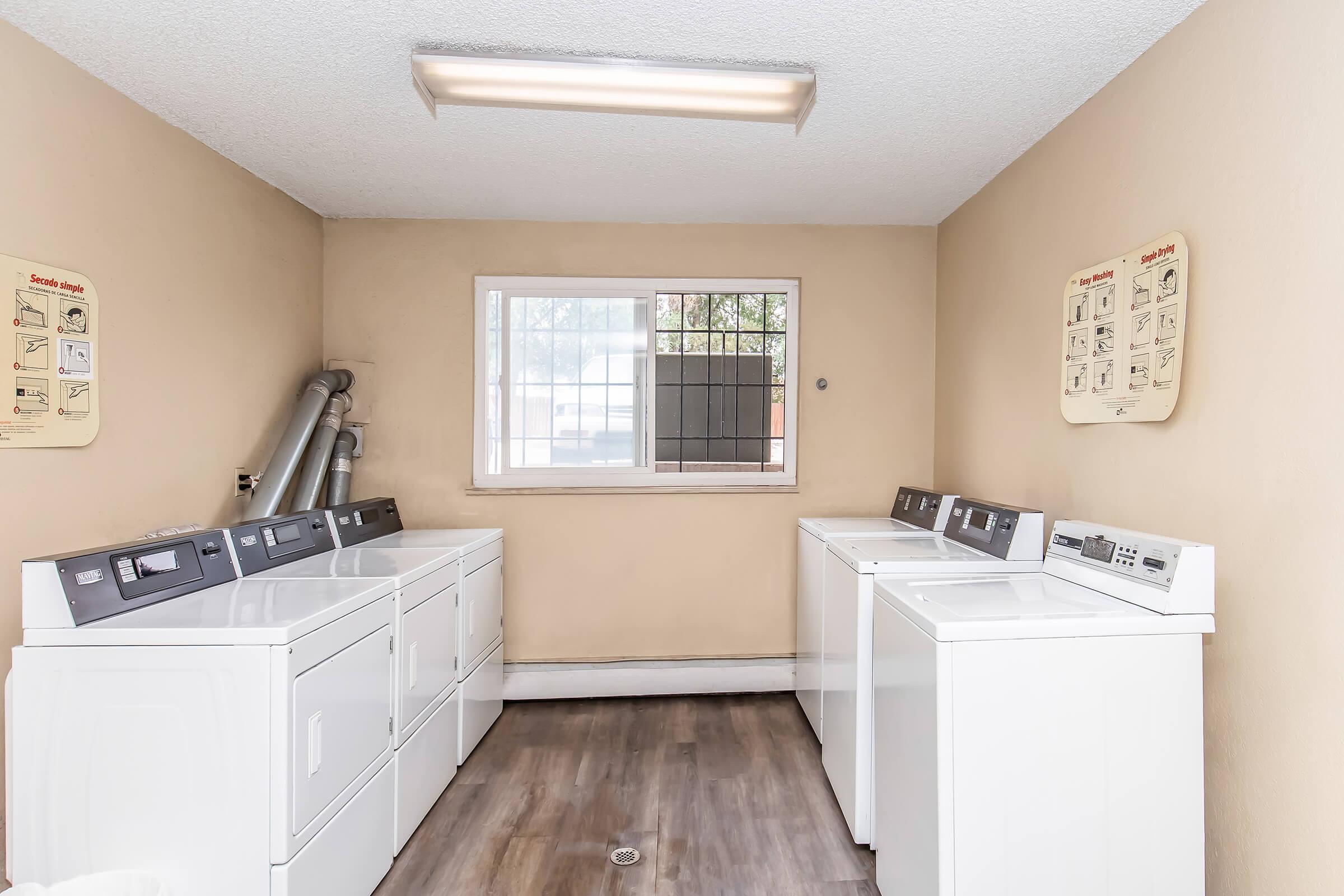 A clean laundry room featuring multiple rows of white washers and dryers against tan walls. Natural light enters through a window, and laundry guidelines are posted on the walls. The floor is wood-like laminate, creating a neat and functional space for doing laundry.