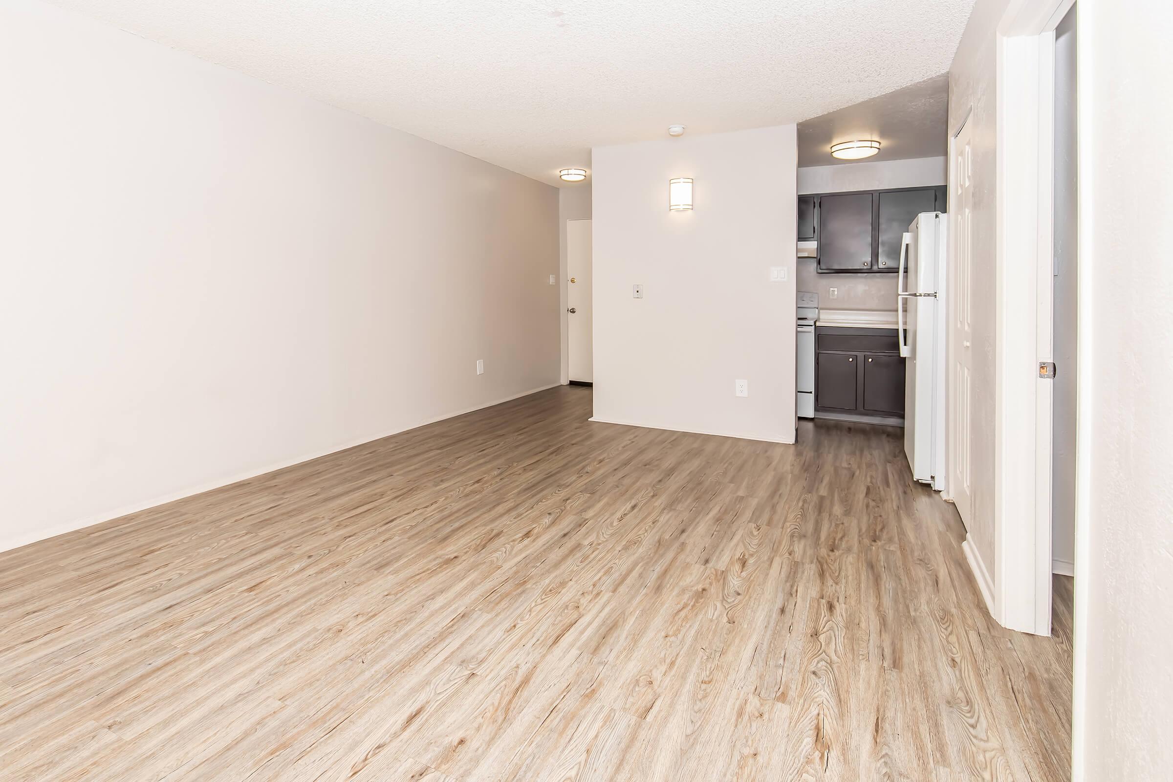 A spacious, empty living area with light-colored walls and wood-style flooring. On one side, a kitchen area with dark cabinetry and white appliances is visible. The room is well-lit and has a neutral aesthetic, creating a bright and open feel.