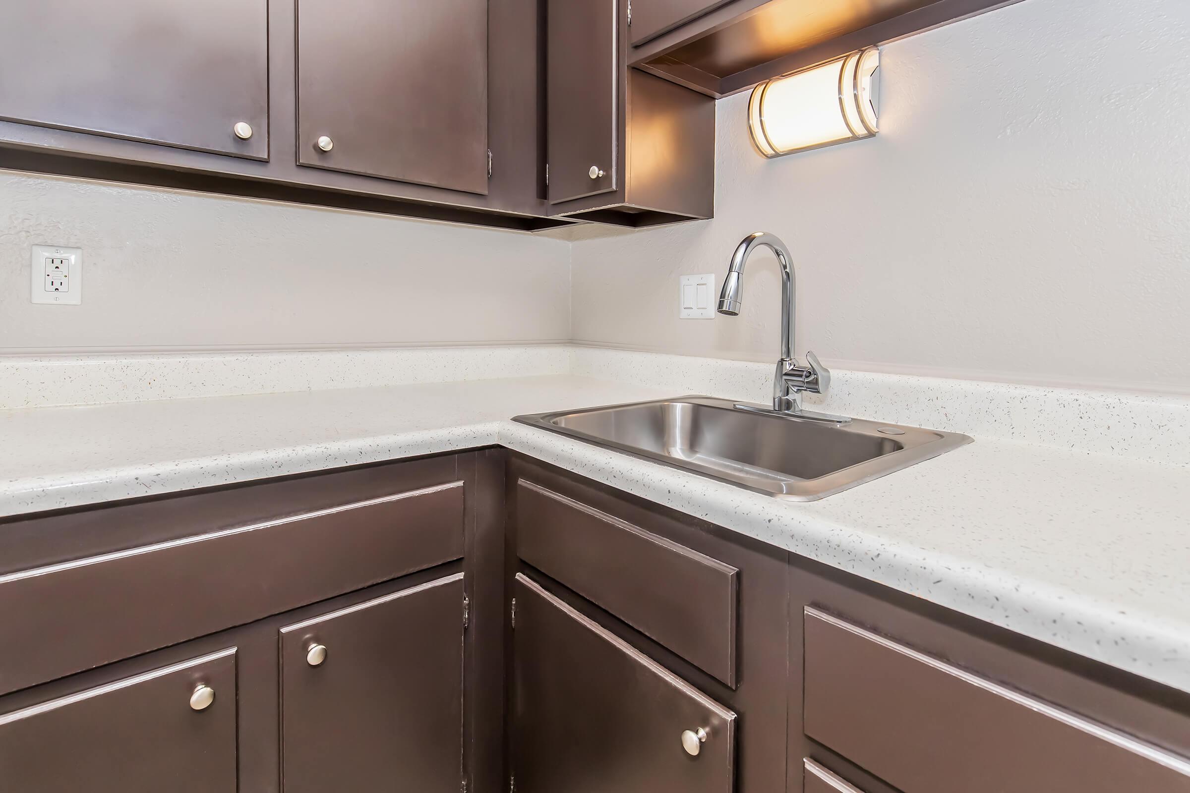 A modern kitchen corner featuring dark wood cabinets, a stainless steel sink, and a light fixture above. The countertop is a light-colored material, and the wall behind is painted a neutral tone. The design is sleek and contemporary, ideal for a functional kitchen space.