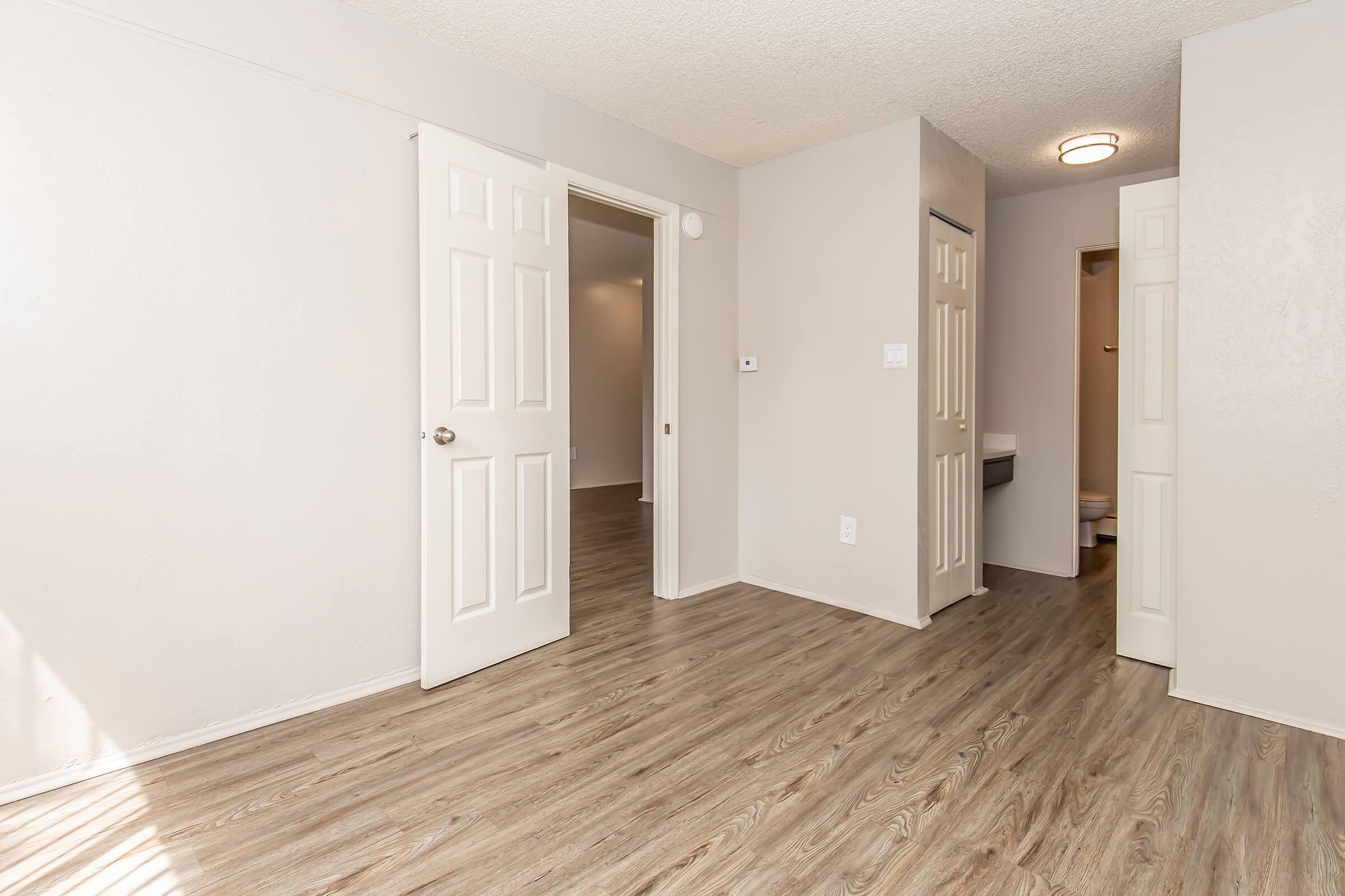 A spacious, empty room featuring beige walls and light wooden flooring. Two open white doors lead to adjacent areas, while a ceiling light fixture provides illumination. The room is well-lit with natural light streaming in, creating a bright and airy atmosphere.