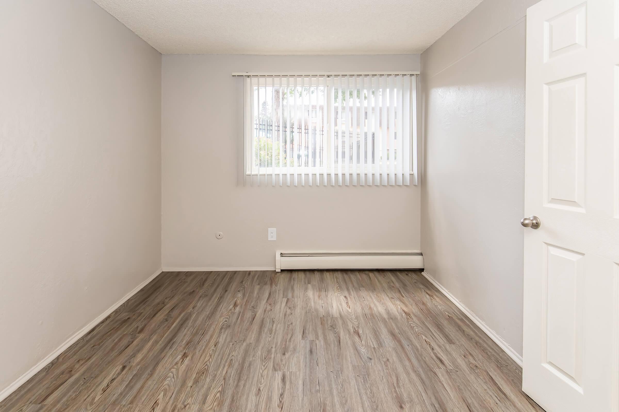 A vacant room with light-colored walls and wood-like flooring. It features a window with vertical blinds allowing natural light, a heating unit beneath the window, and a white door on the right side of the image. The overall atmosphere is bright and minimalistic.