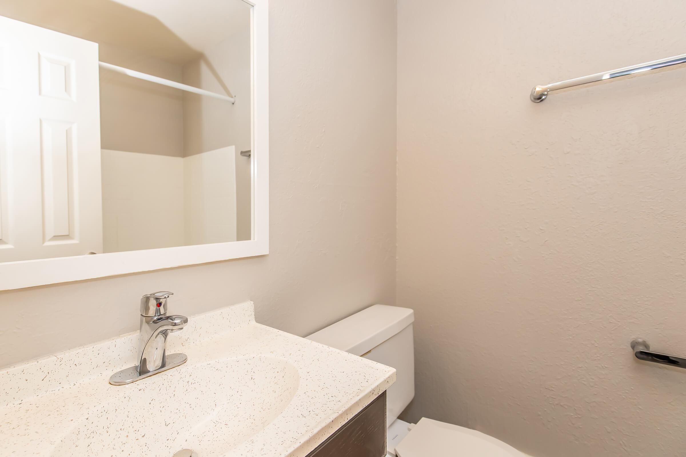 A clean, well-lit bathroom featuring a sink with a modern faucet, a mirror above the sink, a toilet next to a shower stall, and neutral-colored walls. The overall design is simple and functional, with minimal decor.