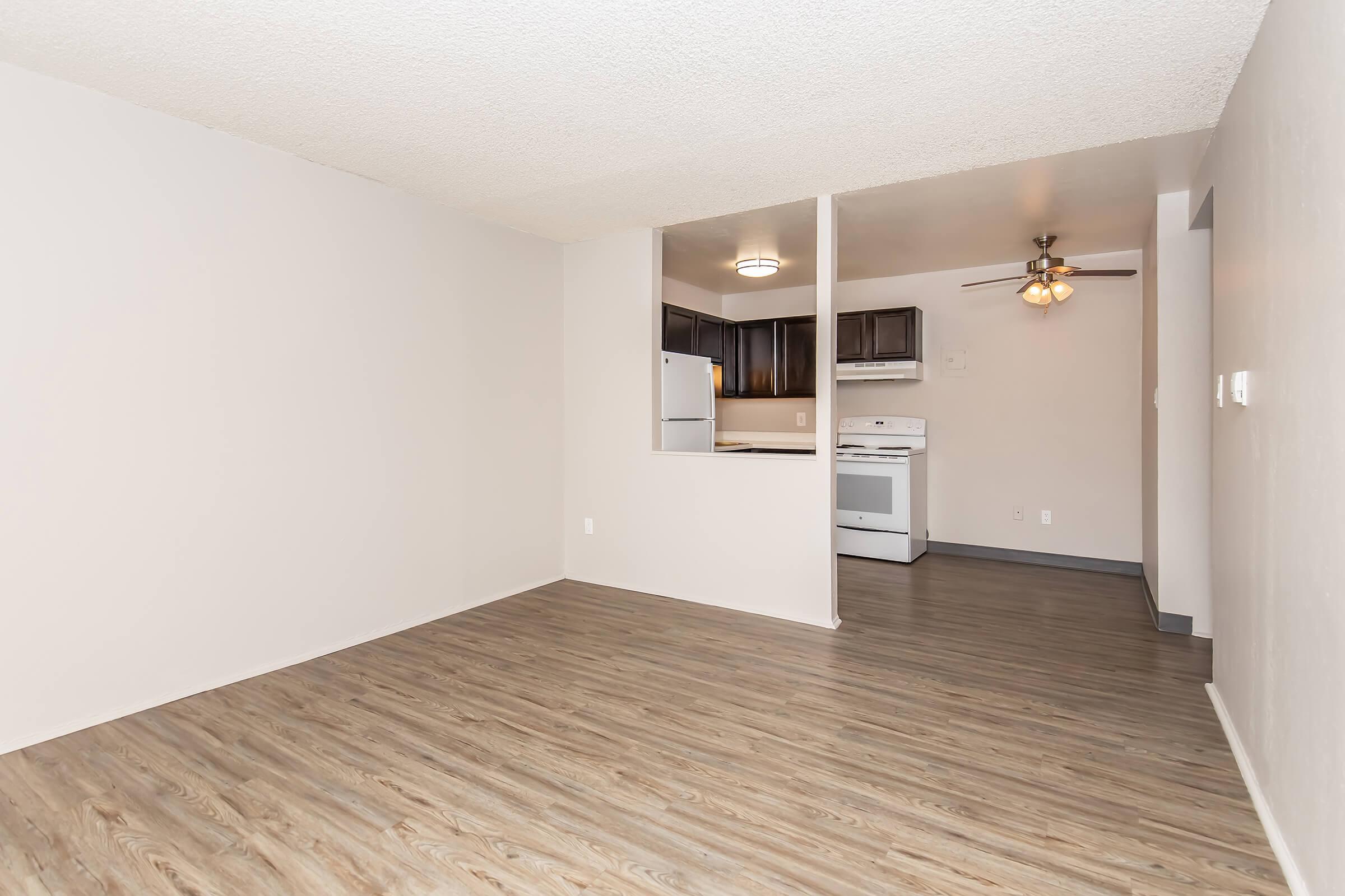 A spacious living area featuring light-colored walls and a wood-style floor. The kitchen is partially visible through an open wall, showcasing dark cabinetry, a refrigerator, and a white stove. A ceiling fan is installed, and the room has ample natural light.