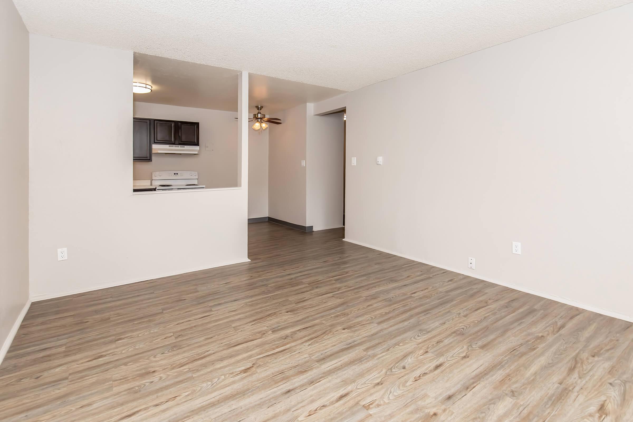 A spacious, modern living room with light-colored walls and a wood-like floor. There is an open layout leading to a kitchen area visible in the background, featuring dark cabinetry and appliances. The room is well-lit and unobstructed, providing a blank canvas for furnishings.