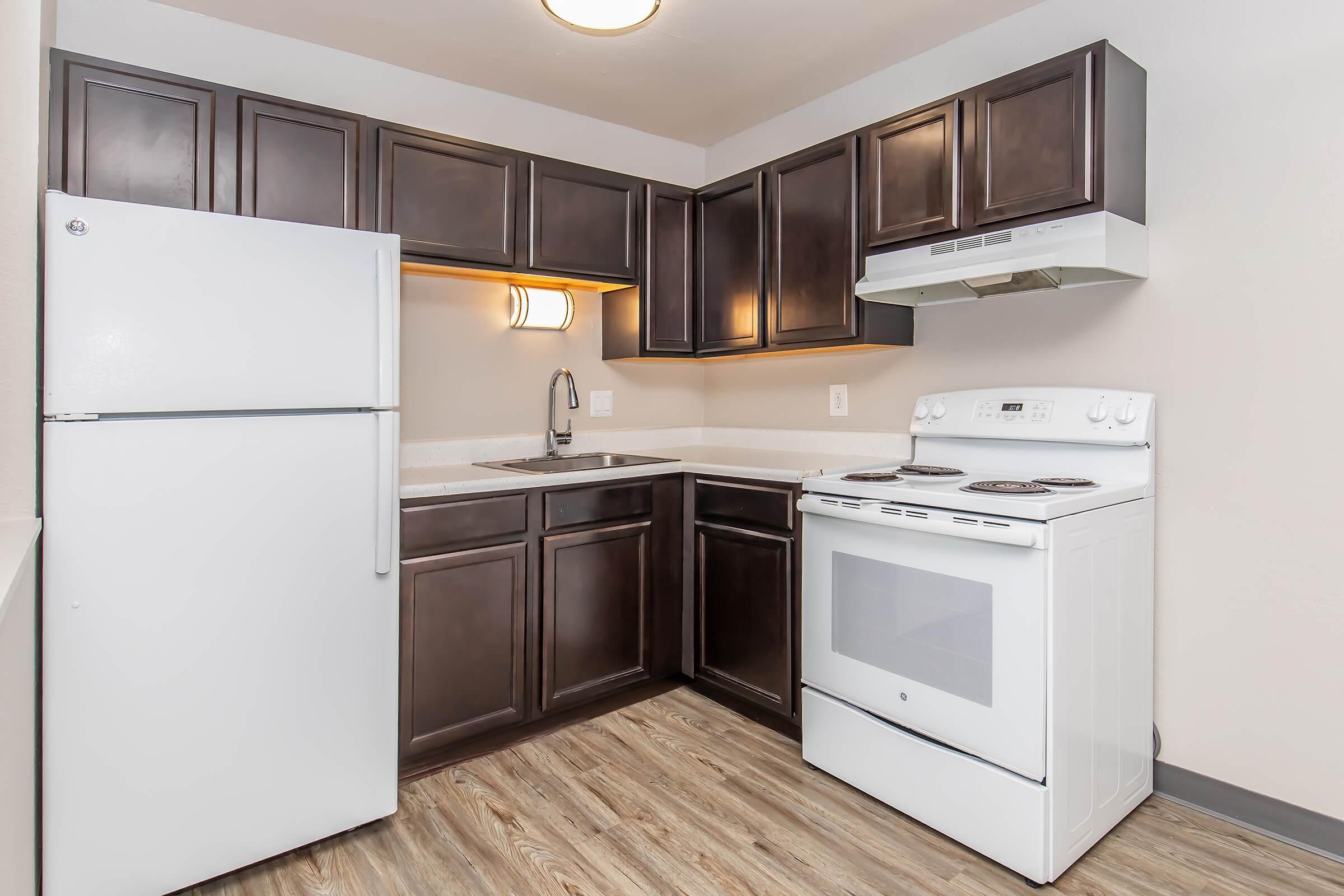 A modern kitchen featuring dark wooden cabinetry, a white refrigerator, a white stove with an oven, and a sink. The countertop is light-colored, and the flooring resembles wood. The kitchen is well-lit with overhead lighting.