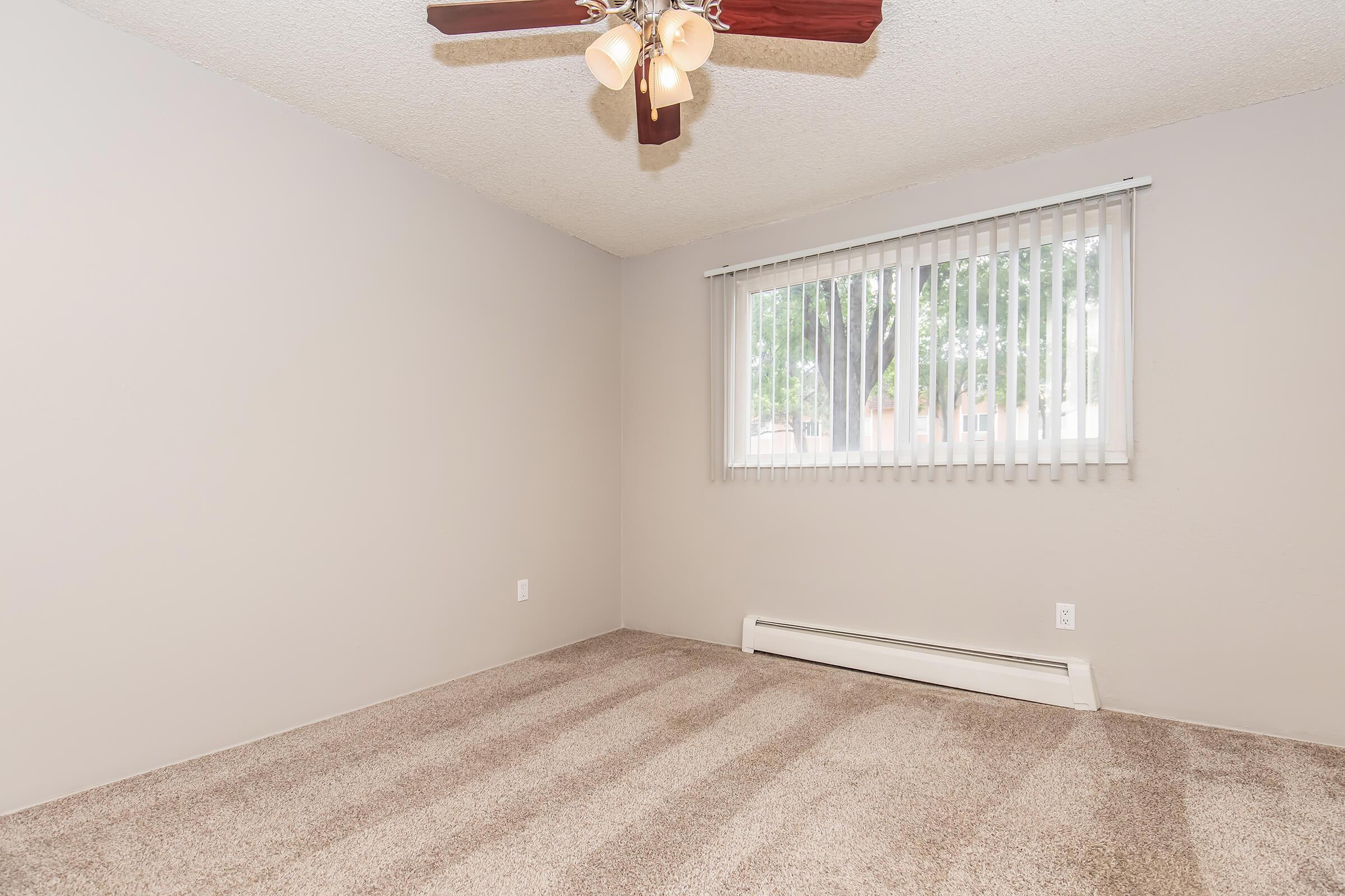 A vacant room featuring light beige walls, a ceiling fan with three bulbs, and sliding vertical blinds on a window. The floor is covered with light beige carpet, and there is a baseboard heater visible along the wall. The space is bright and uncluttered, ideal for personal decoration.