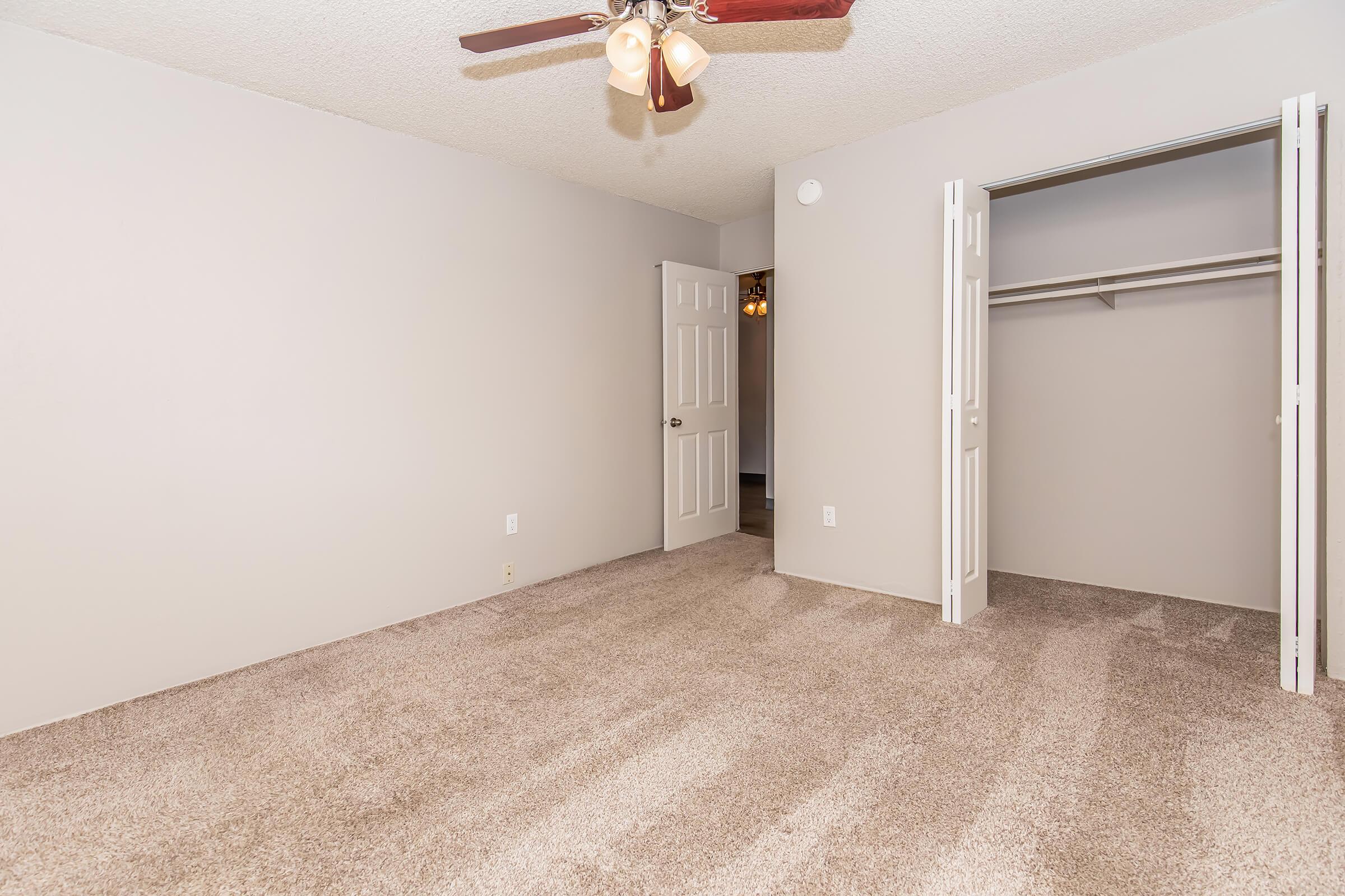 A clean, empty room with light gray walls and a ceiling fan. The floor is covered in beige carpet. On one side, there are two open white closet doors, and a doorway leads to another area. The room appears spacious and well-lit, suitable for various uses.