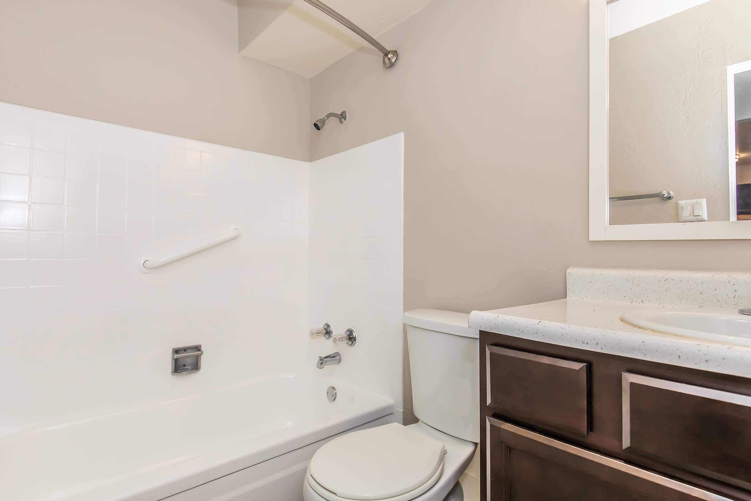 A clean, modern bathroom featuring a white bathtub with a showerhead, a dark wood vanity with a sink, and a toilet. The walls are painted a neutral color, and there is a mirror above the sink. The overall design is simple and contemporary.