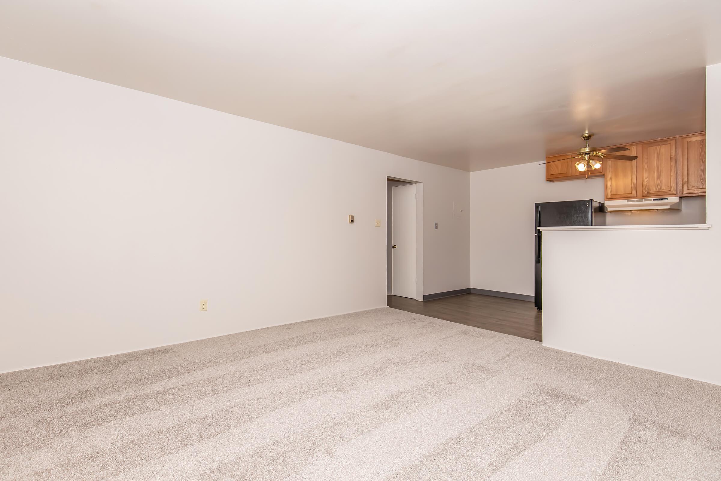 A spacious, empty room with light-colored walls and a carpeted floor. In the background, there's a kitchen area with wooden cabinets, a ceiling fan, and a black refrigerator. The doorway leads to another room, creating a simple, clean layout ideal for various uses.