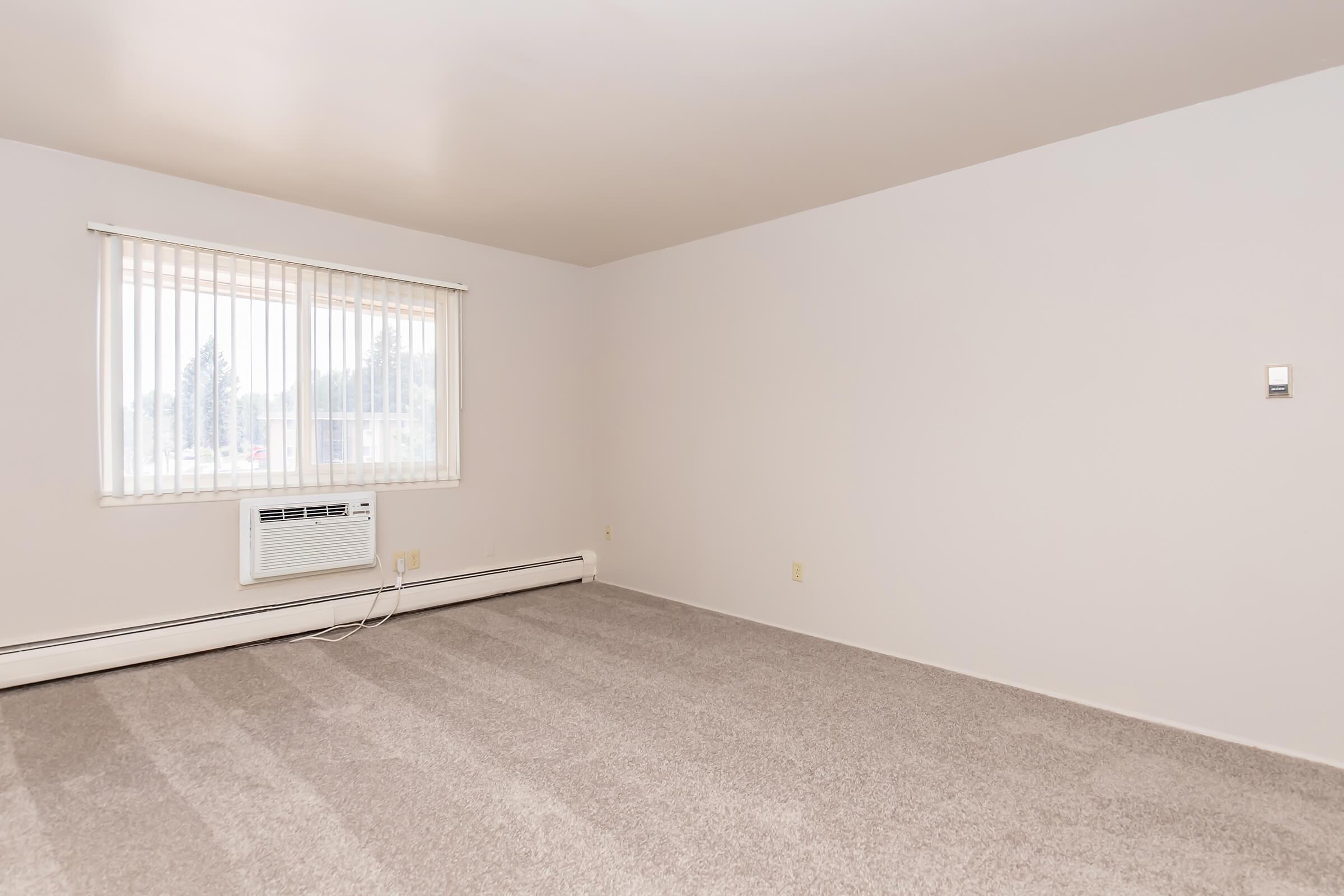 A spacious, empty room featuring light gray carpet and a large window with white blinds. An air conditioning unit is mounted beneath the window, and the walls are painted white. The room is well-lit, emphasizing its open layout and potential for furnishing.