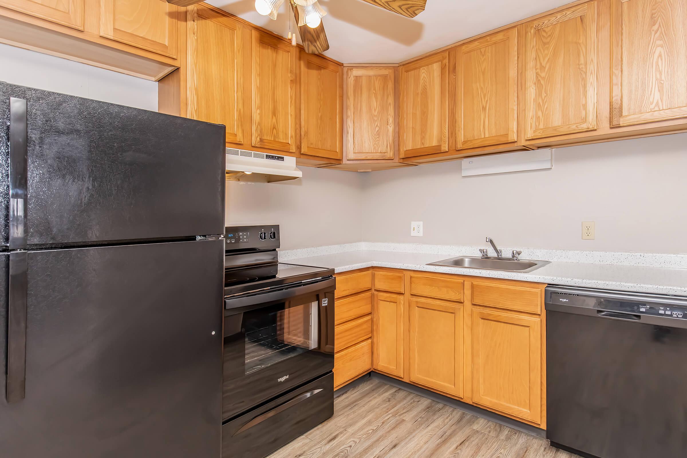A modern kitchen featuring dark appliances, including a refrigerator and oven, set against light granite countertops. Wooden cabinets provide storage space, while a small sink is visible. The flooring is a stylish light wood. A ceiling fan is installed above for ventilation.
