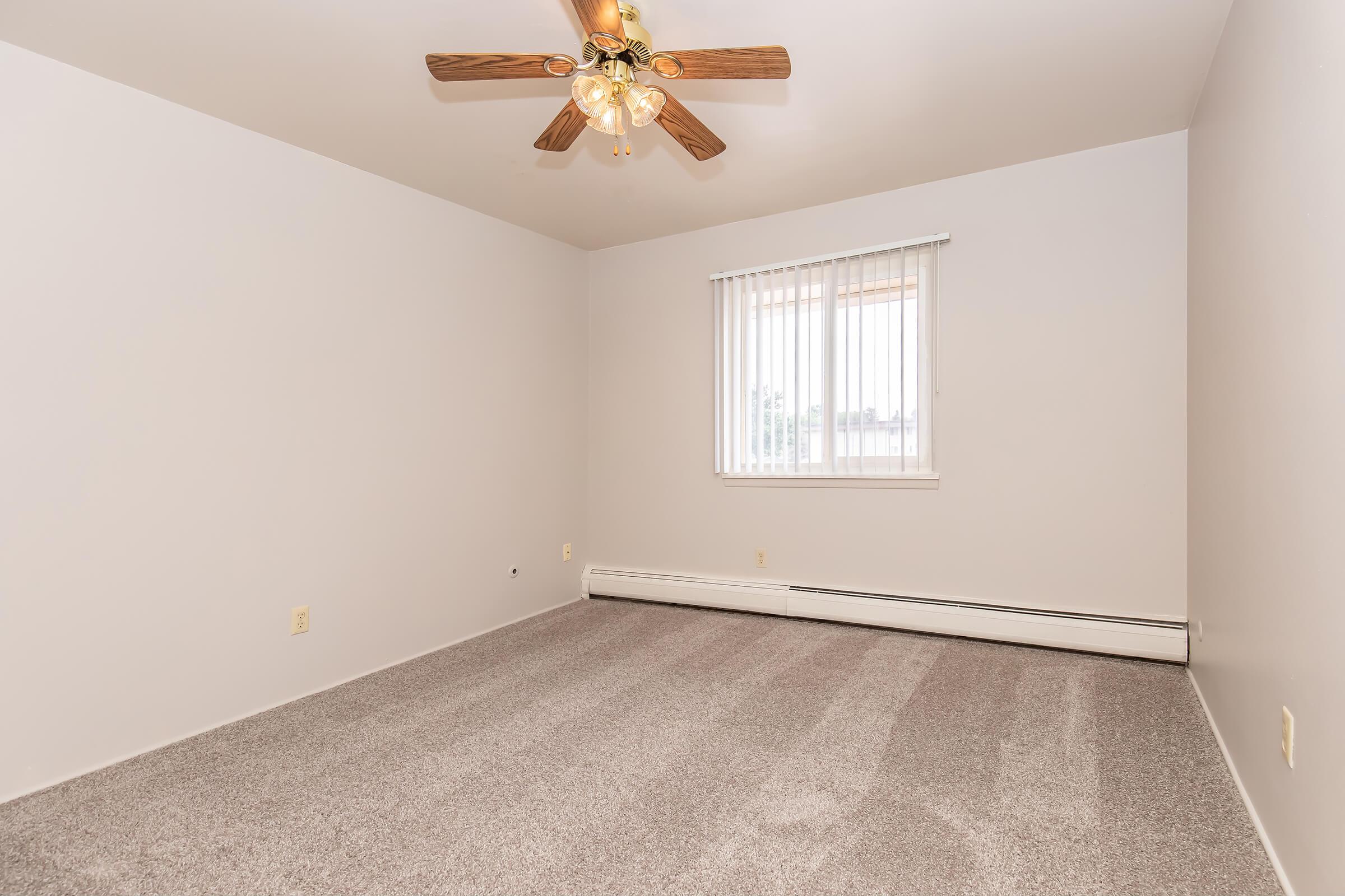 Empty room with light gray walls and carpet. A ceiling fan with wooden blades hangs from the ceiling. There is a window with vertical blinds allowing natural light to enter. No furniture or decorations are present, giving the space a clean and minimalist appearance.