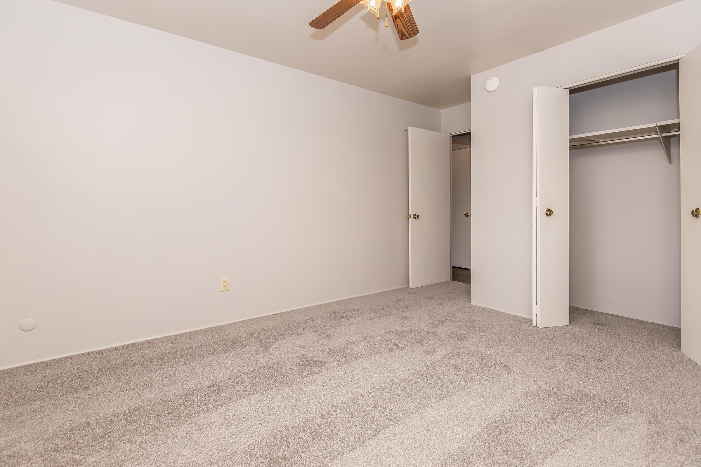 Empty room with light gray walls and carpeted floor. A ceiling fan is overhead, and there are two closed white doors on the left and right sides. A closet is visible beyond one of the doors, adding to the spacious feel of the room. Natural light creates a bright atmosphere.