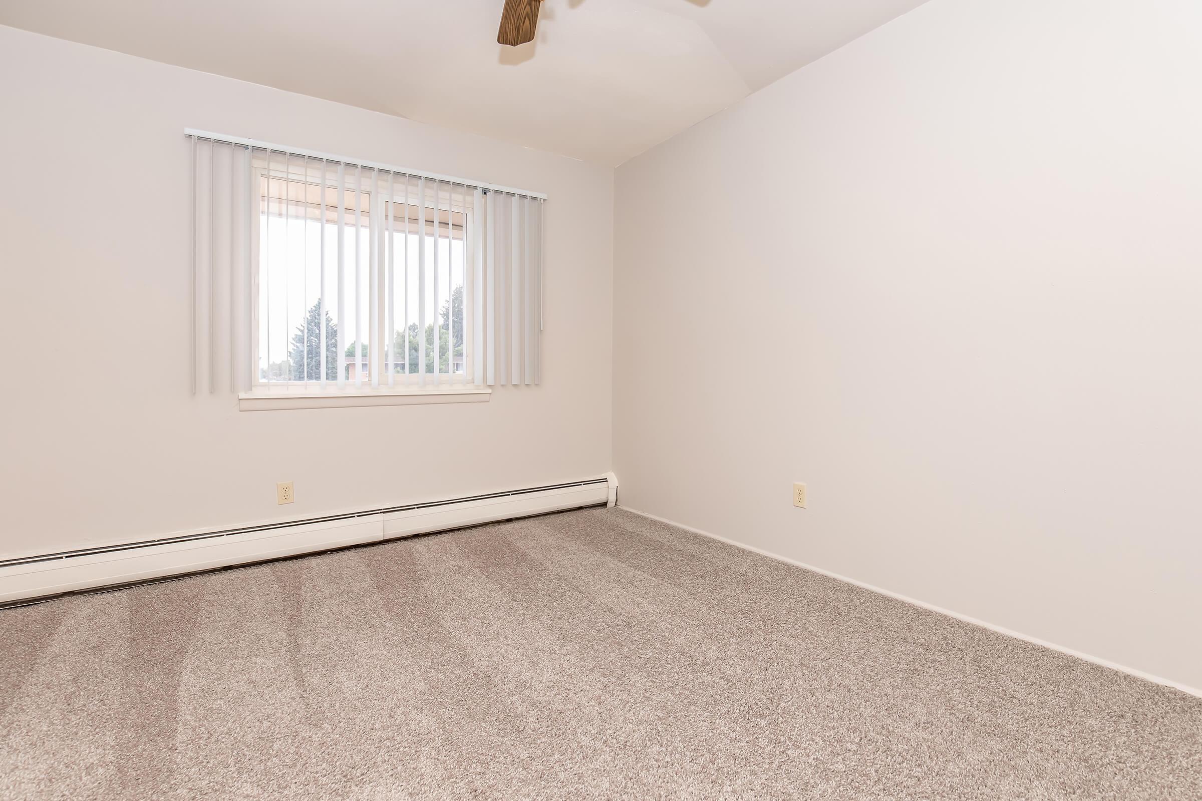 Empty room with light gray carpet, white walls, and a window featuring vertical blinds. There is a ceiling fan and baseboard heating along the wall. The space is well-lit by natural light coming through the window, creating an open and airy atmosphere. Ideal for various uses or personalization.