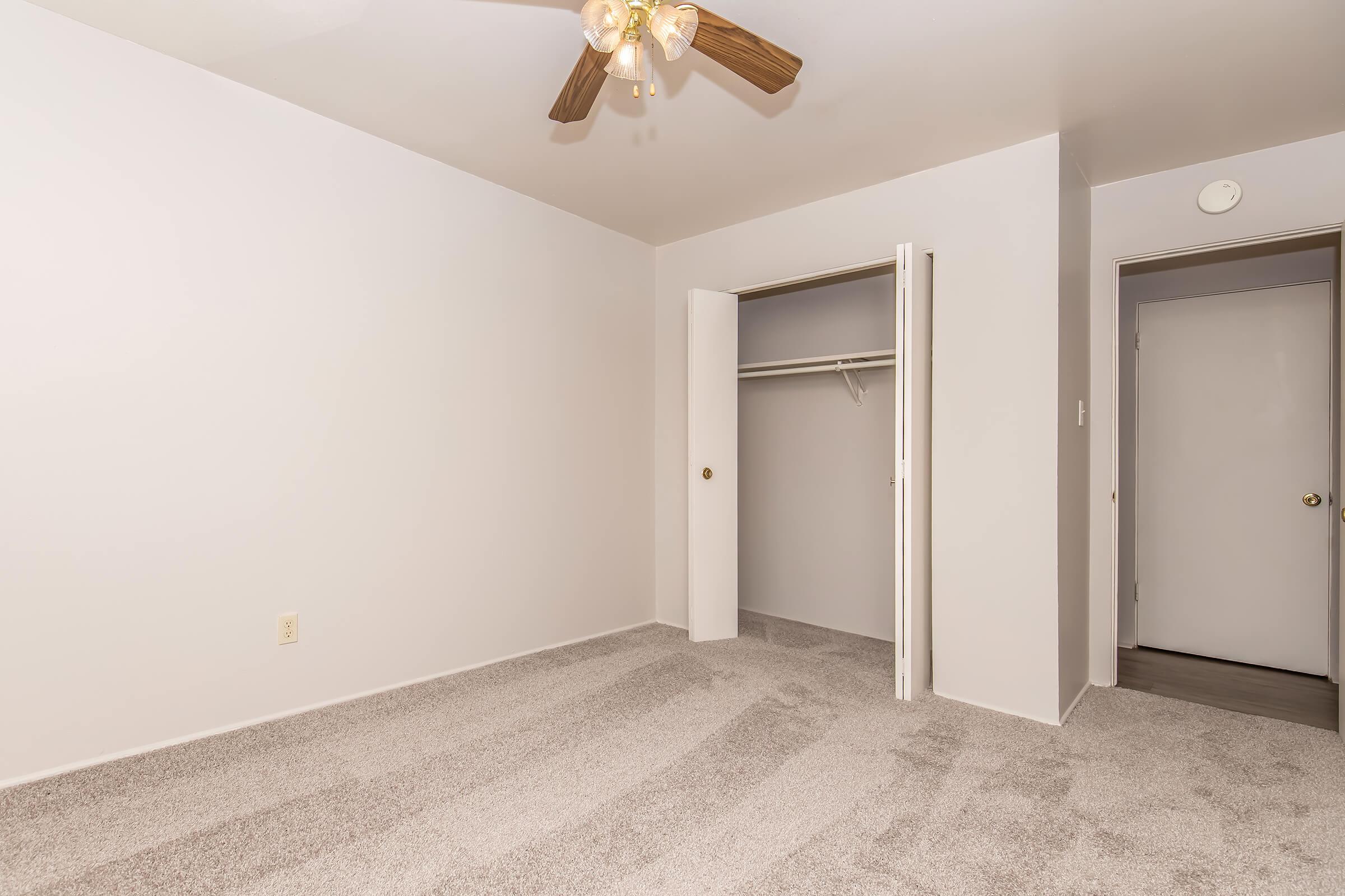 Empty room with light-colored walls, a ceiling fan with wooden blades, and freshly carpeted flooring. There is a closet with an open door on the left and a closed door on the right leading to another space. Natural light brightens the room, creating a clean and spacious atmosphere.