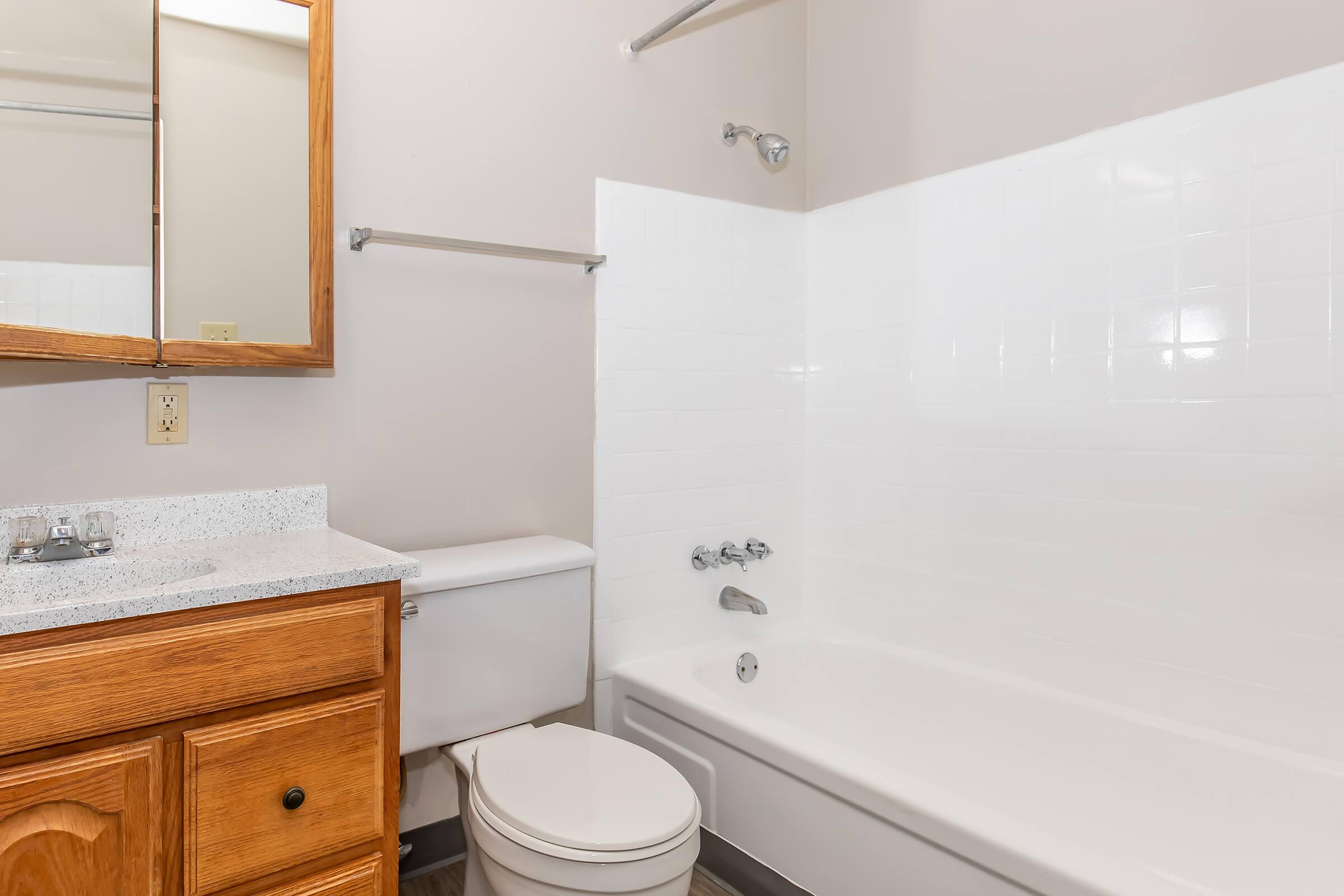 A clean, modern bathroom featuring a white bathtub with a showerhead, a wooden cabinet with a sink, a toilet, and a large mirror. The walls are painted light gray, and the flooring is a subtle dark material. The overall design is simple and functional.
