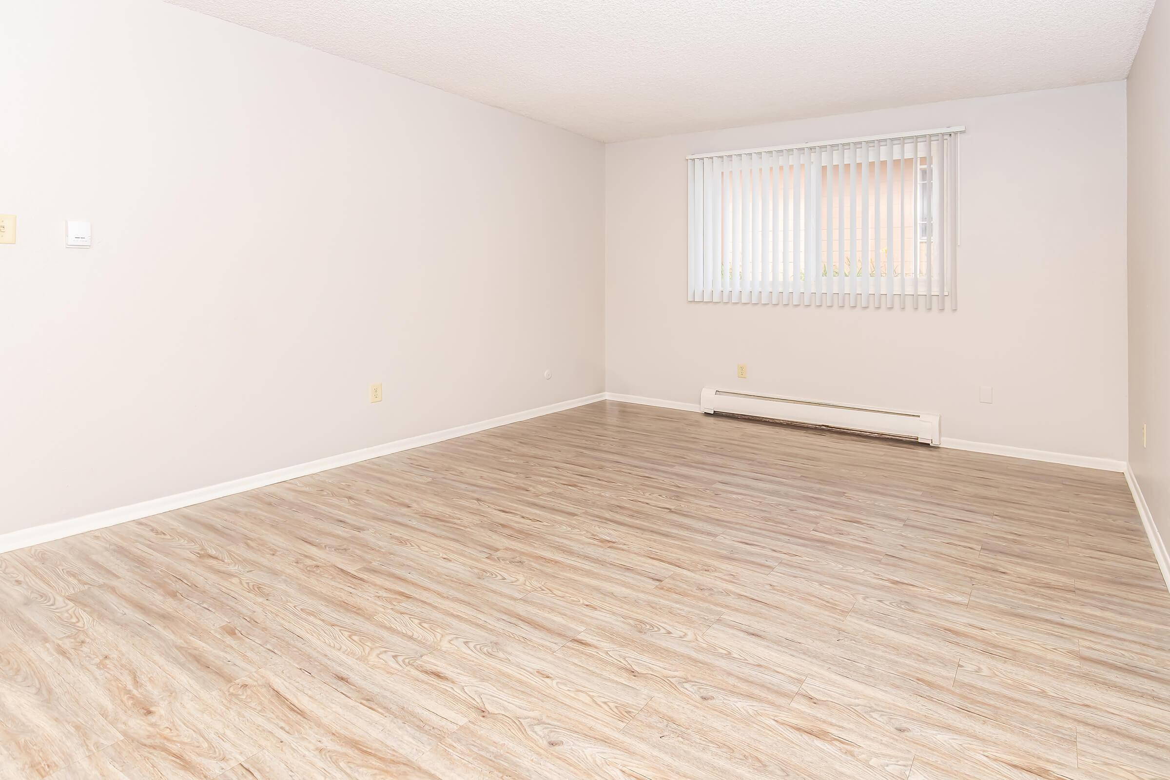 A sparsely furnished room with a light-colored wood floor, beige walls, and a window covered by vertical blinds. The space is empty, creating a clean and open atmosphere, ideal for various uses.