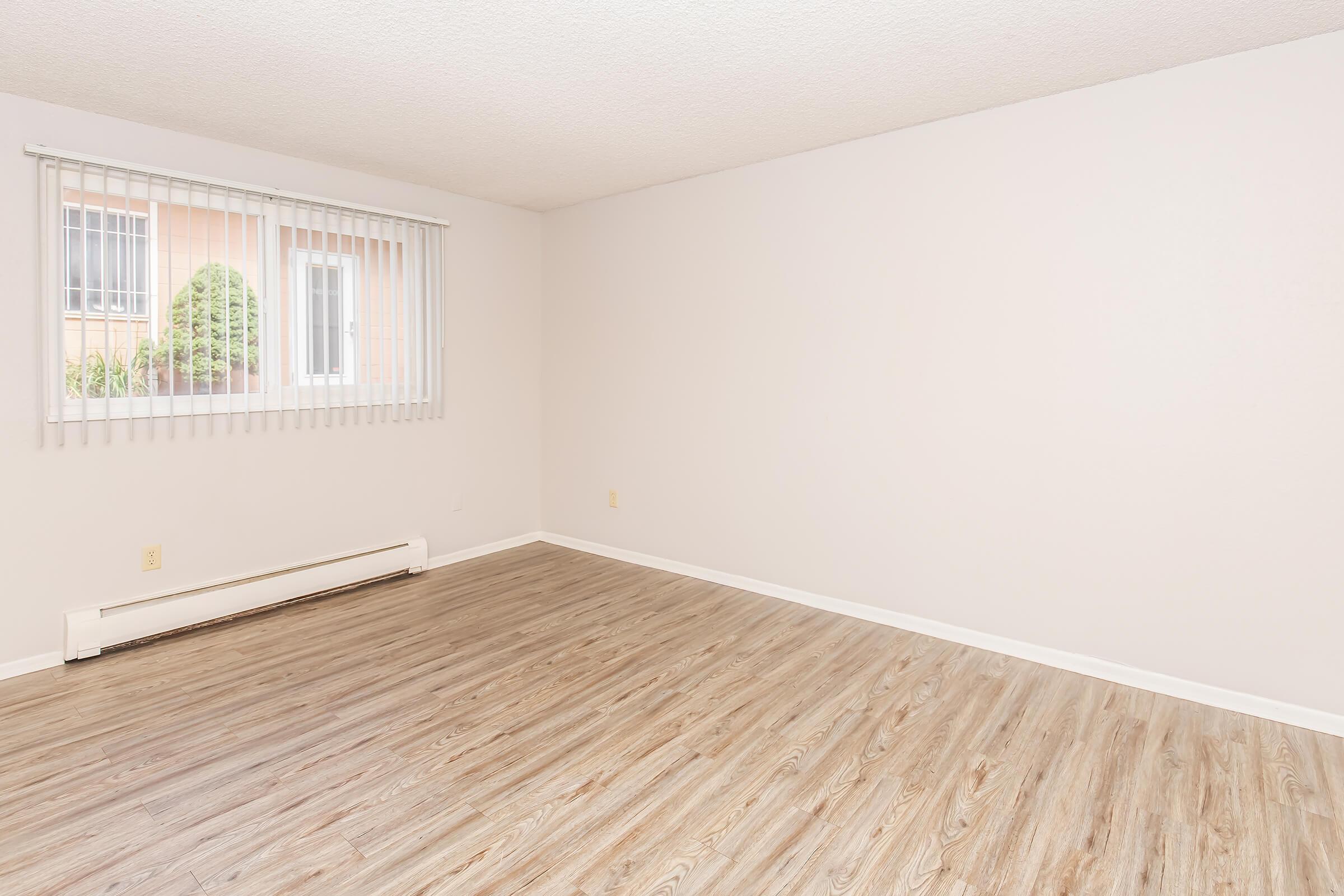 A spacious, empty room with light-colored walls and a wooden floor. A window with vertical blinds allows natural light to enter. The room has a simple, uncluttered appearance, suitable for various uses such as living space or office.