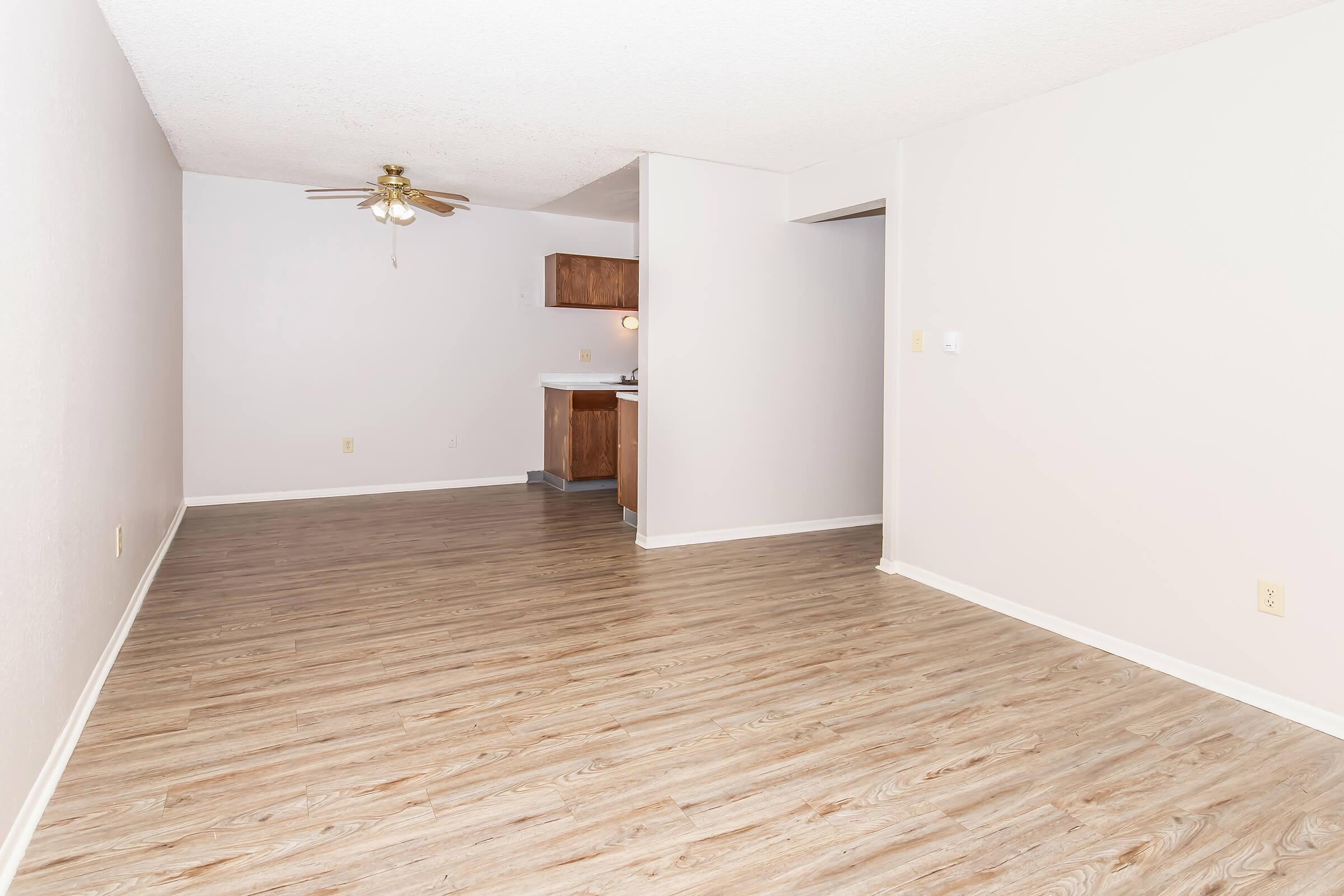 A spacious, empty living room with light-colored walls and a ceiling fan. The floor features wooden laminate, and a small kitchenette is visible in the background. The overall atmosphere is bright and inviting, perfect for new furnishings and decor.