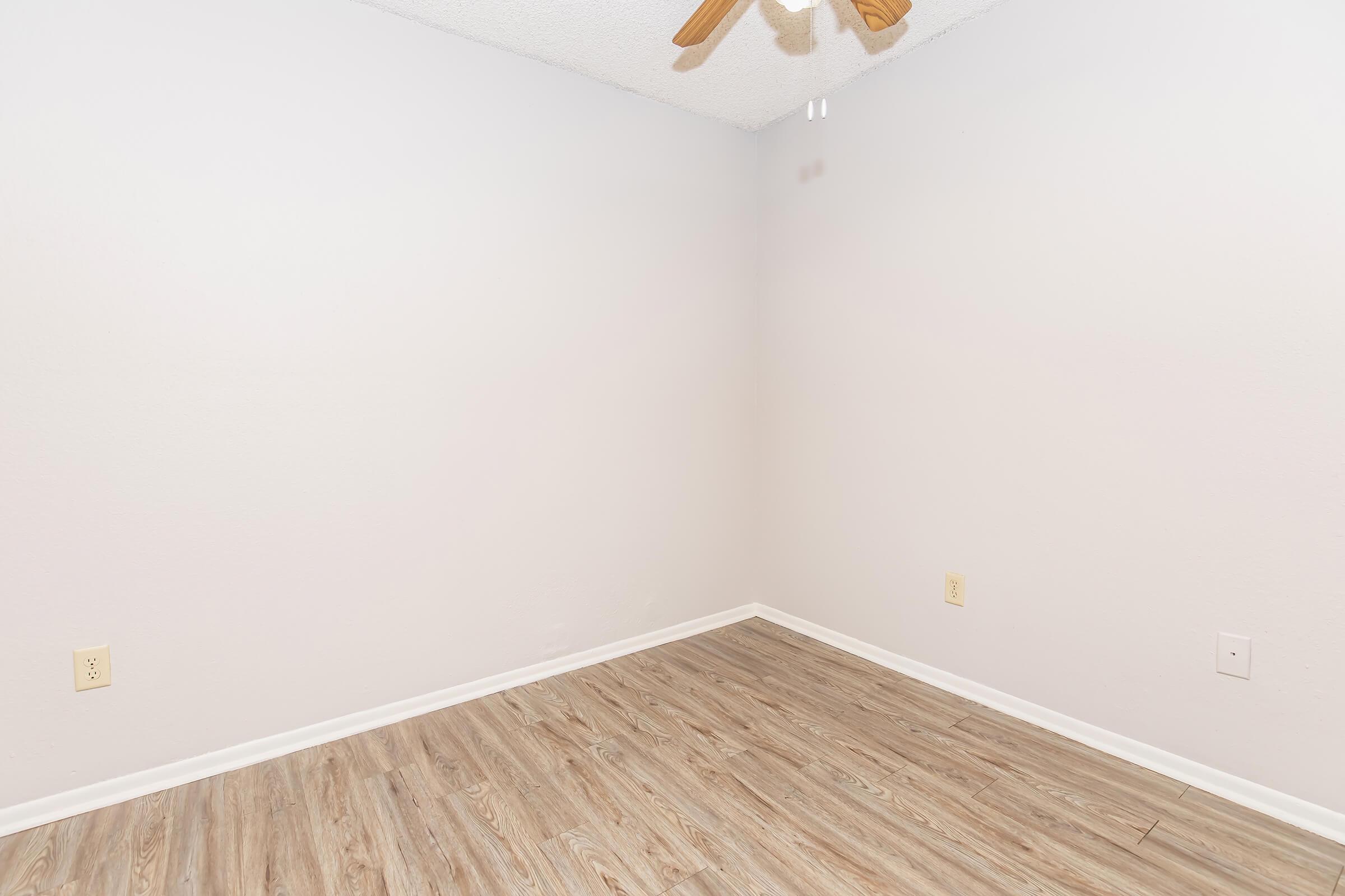 A sparsely furnished room with light gray walls and a wooden floor. The space features a ceiling fan, a couple of electrical outlets, and no furniture or decorations. The overall ambiance is minimalistic and neutral, creating a blank canvas for various uses.
