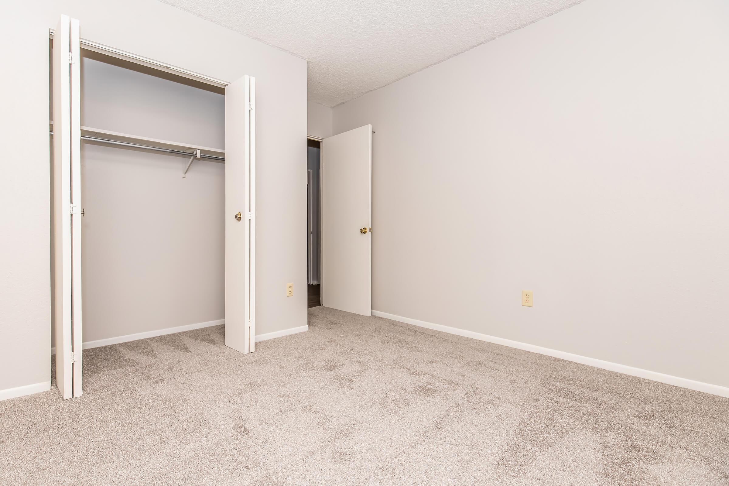 A clean, empty room with light gray walls and a plush carpet. One wall features two opened closet doors, revealing a simple, undecorated closet space. A closed white door is visible on the right, leading to another area. The overall look is minimalistic and spacious, inviting for potential decoration or use.