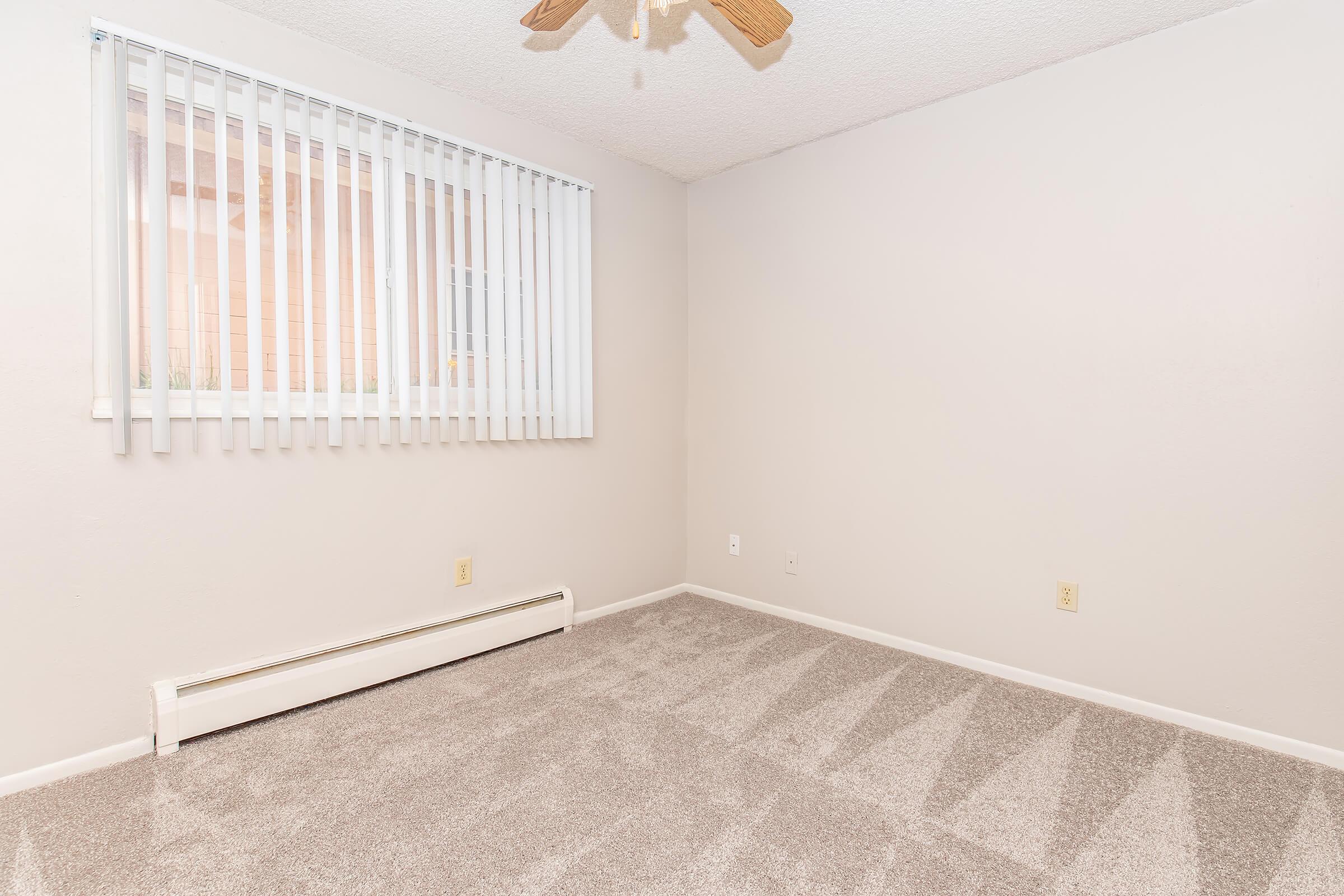A light and neutral-colored empty room with beige walls and a gray carpet. There's a small window with vertical blinds allowing natural light, and a ceiling fan above. The space appears clean and ready for furnishing, with no visible decor or items present.