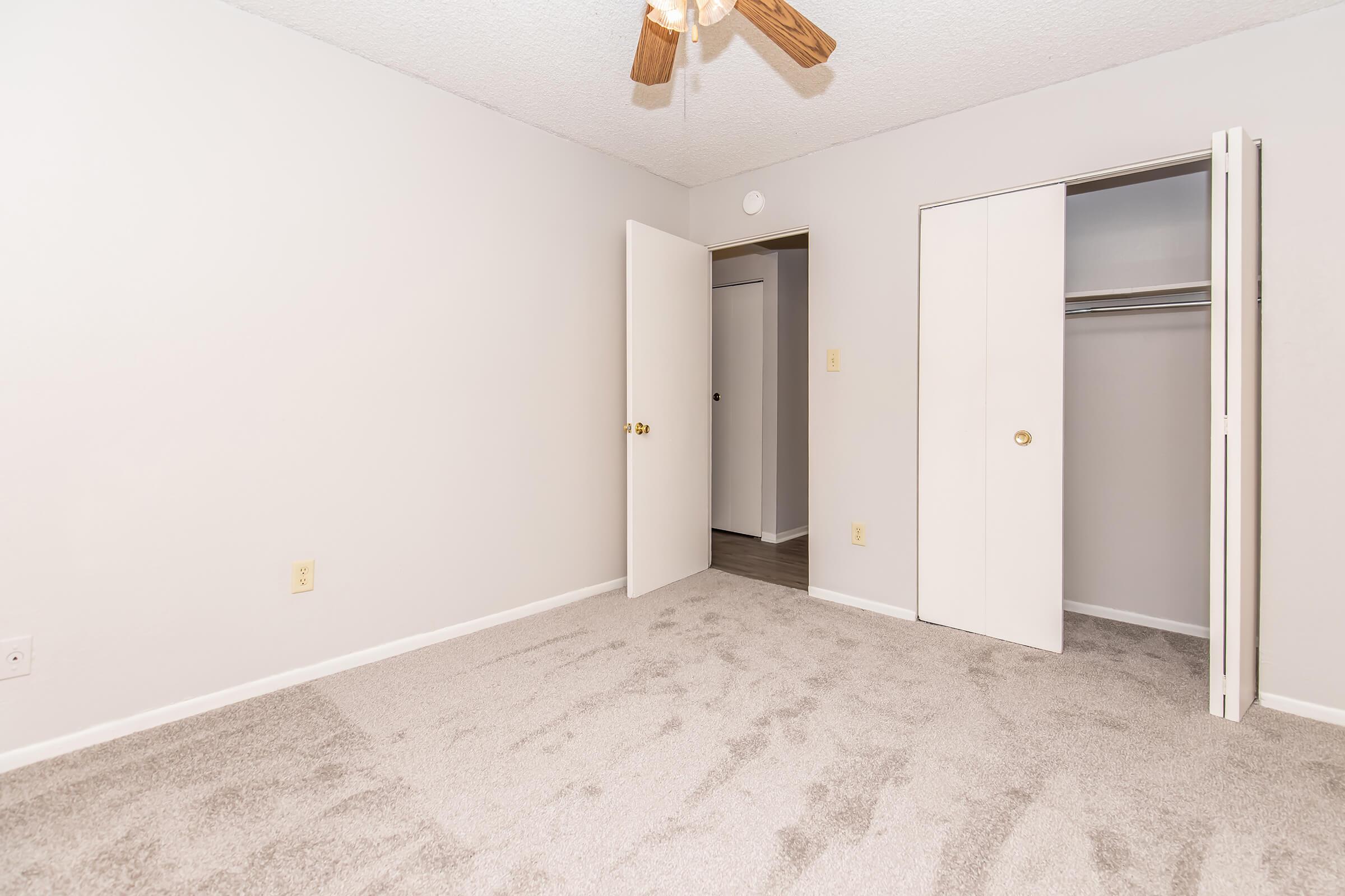 A sparsely furnished bedroom with light-colored walls and a ceiling fan. The floor is covered with beige carpet. There are two doors: one leads to a closet, while the other opens to a hallway. Natural light illuminates the room, creating a bright and airy atmosphere.