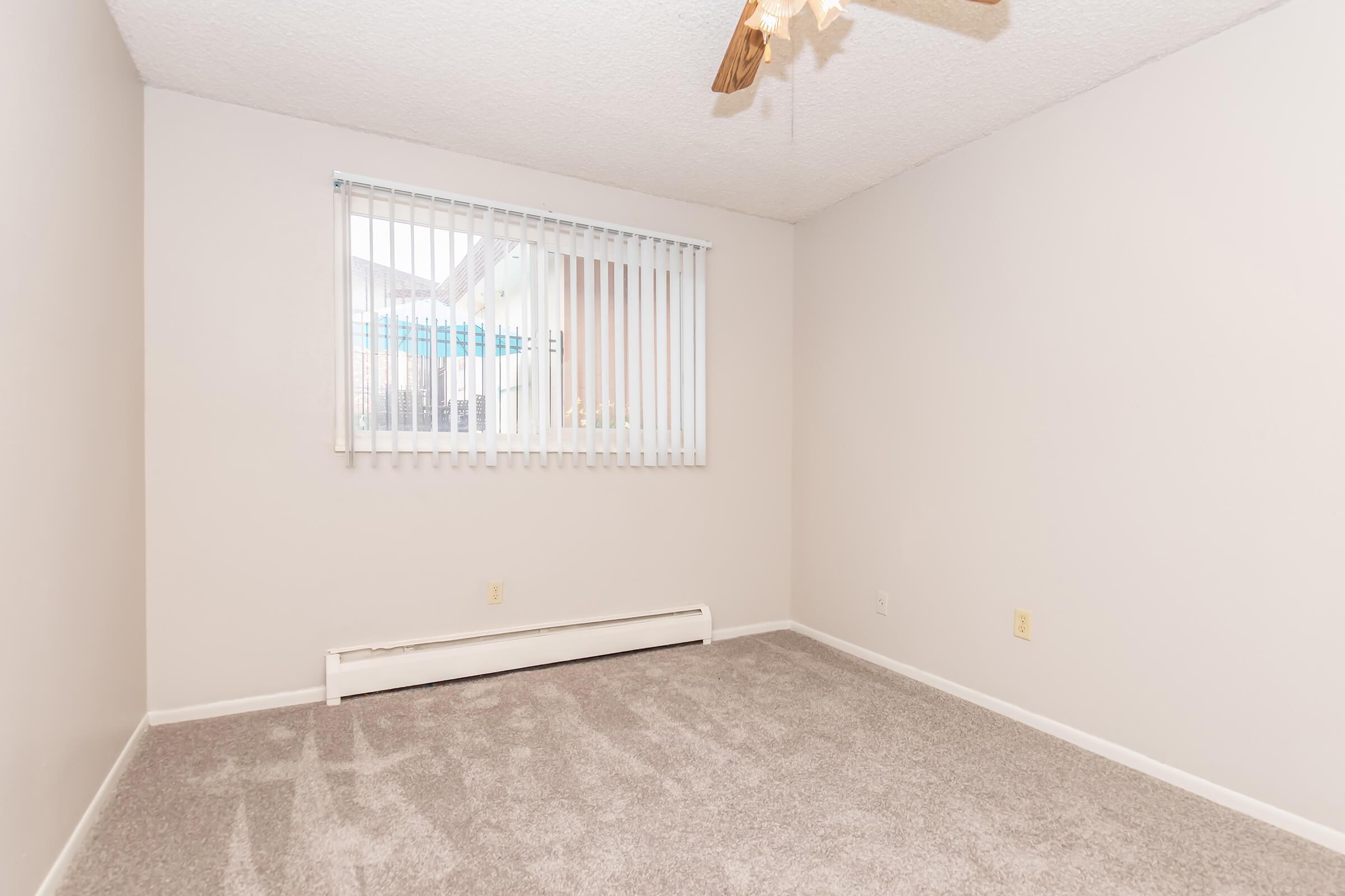 Empty room with light beige walls and a light gray carpet. A window with vertical blinds allows natural light to enter. A ceiling fan is visible, but there is no furniture or decoration in the space, creating a clean and open atmosphere. Heating unit is located along the wall beneath the window.