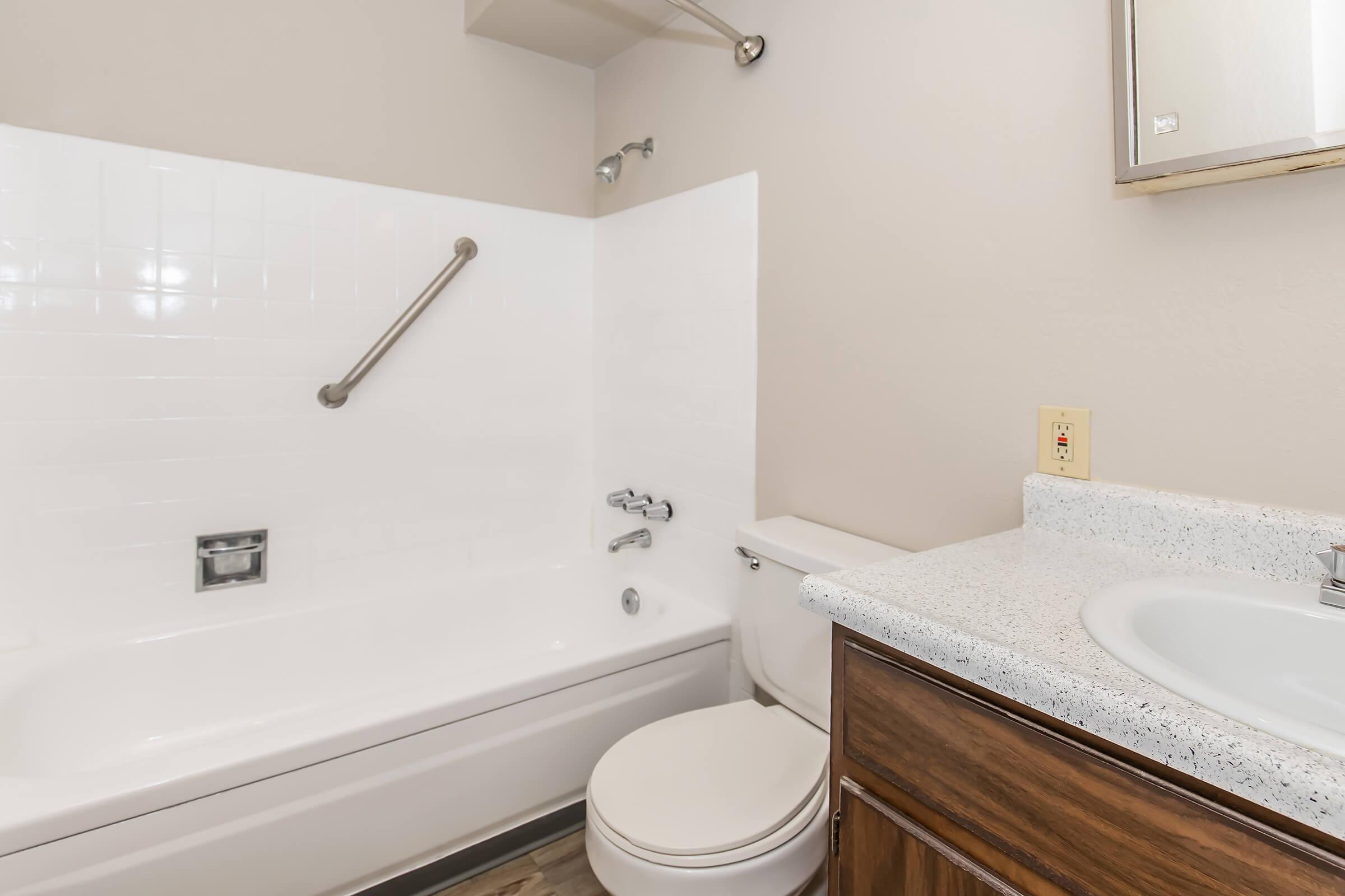 A clean and well-lit bathroom featuring a white bathtub with a handrail, a toilet, and a sink with a granite countertop. The walls are light beige, and there is a wall-mounted mirror above the sink. The flooring is made of wooden planks, enhancing the space's warmth and modern look.