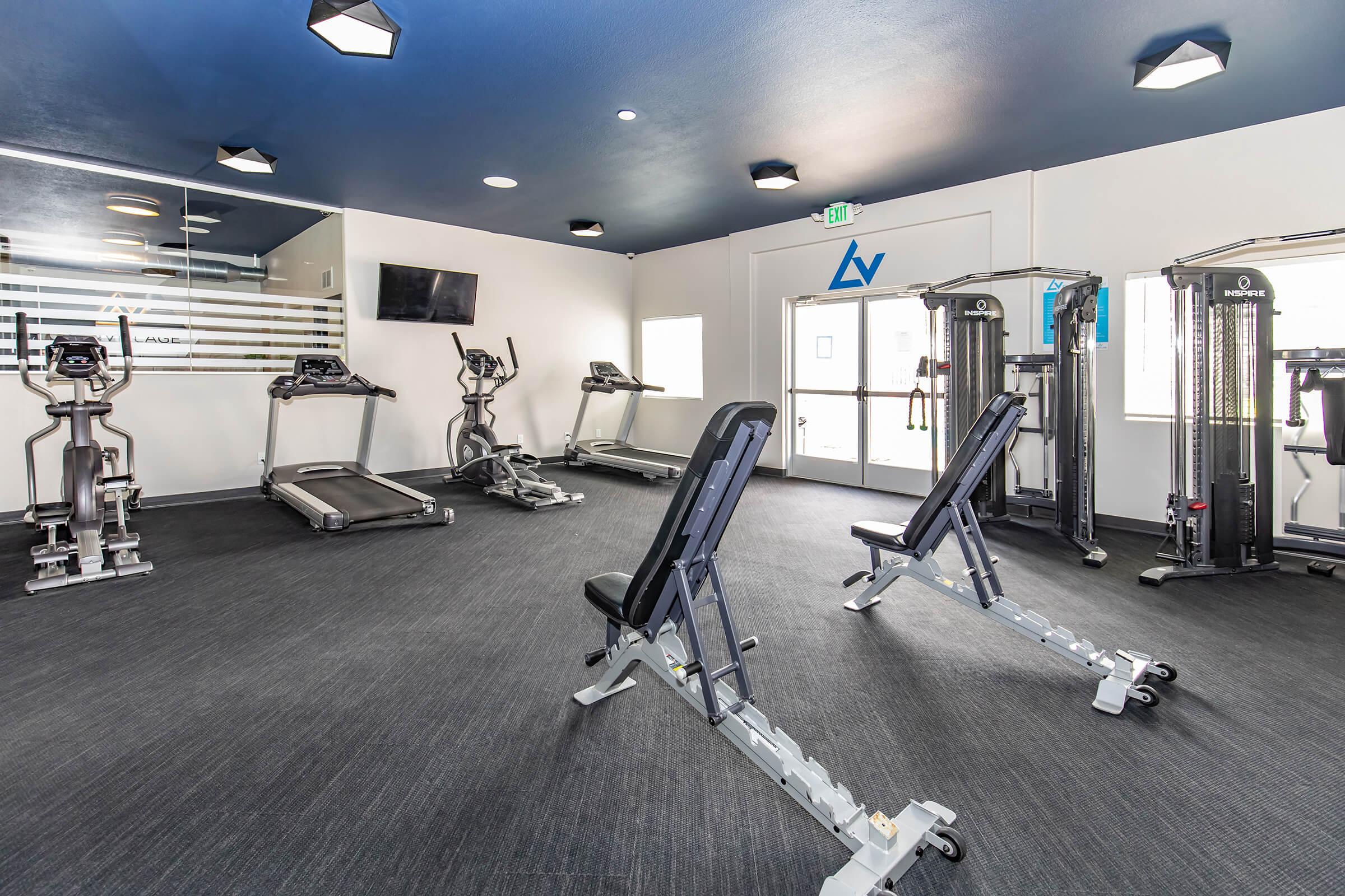 A modern gym interior featuring multiple exercise machines, including treadmills and weight training equipment. The space is well-lit with a blue ceiling and a wall-mounted TV. Large windows provide natural light, and the floor is sleek and dark.