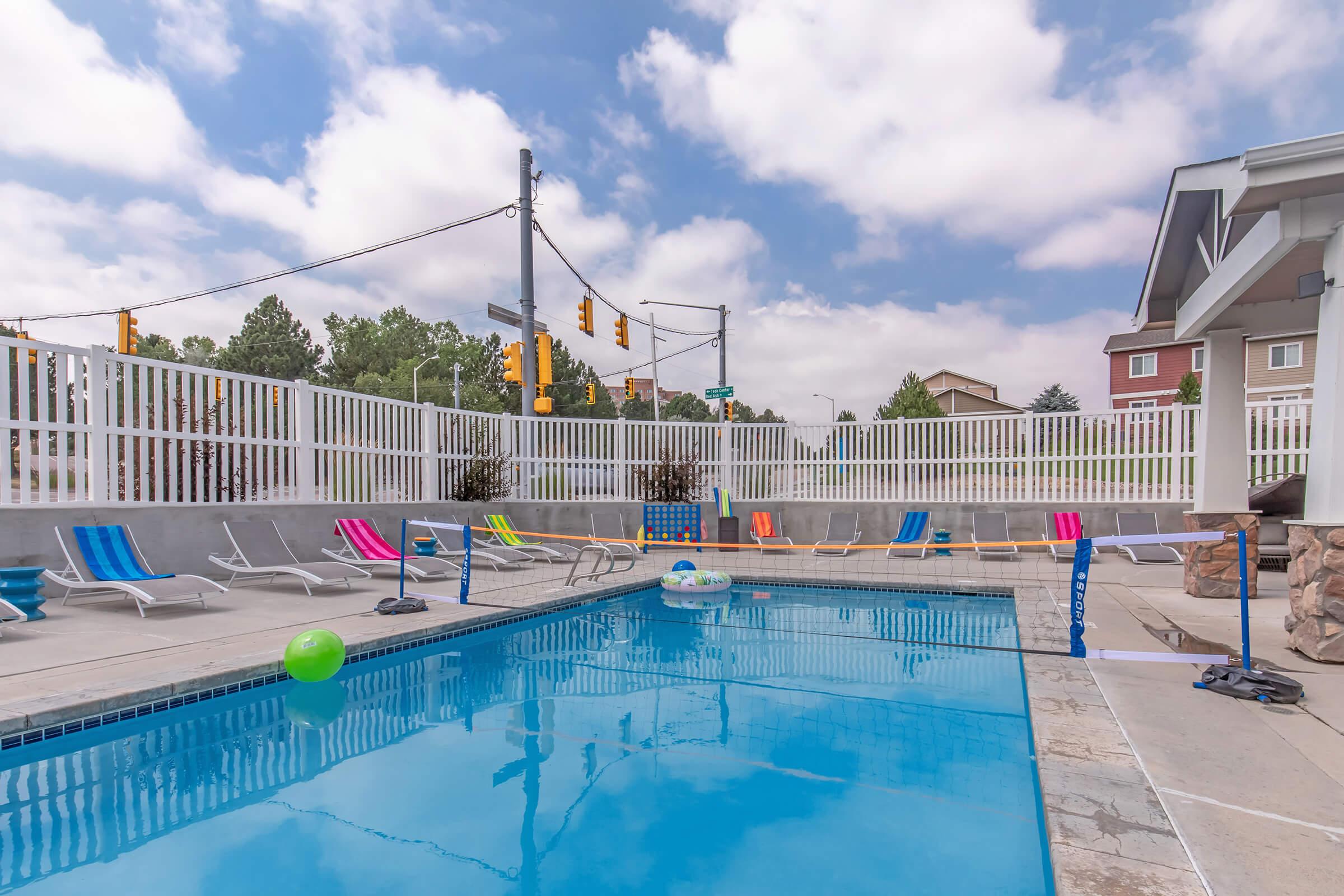 A clear swimming pool surrounded by lounge chairs and colorful floaties. The area features a white picket fence and is set against a backdrop of a cloudy blue sky and nearby buildings. Traffic lights can be seen in the background.