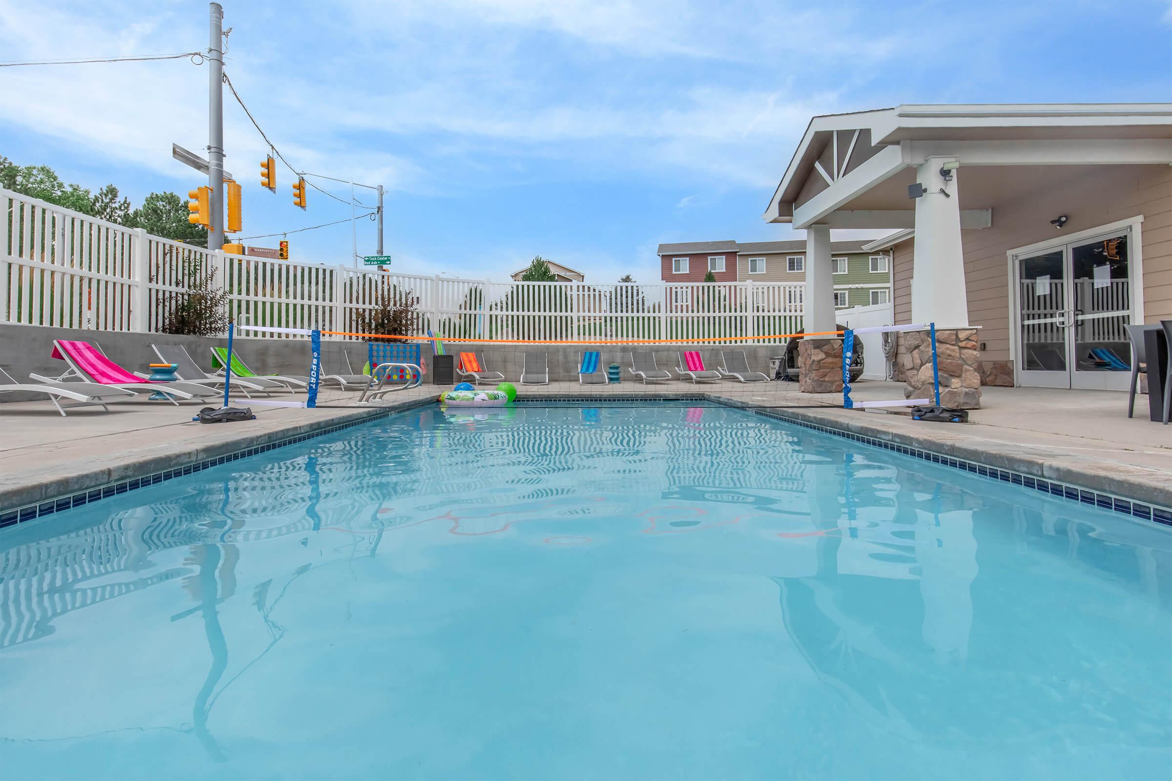 A clear swimming pool surrounded by lounge chairs and colorful pool floats, with a white fence in the background. The area is well-lit and features a blue sky overhead, creating a relaxing outdoor environment.