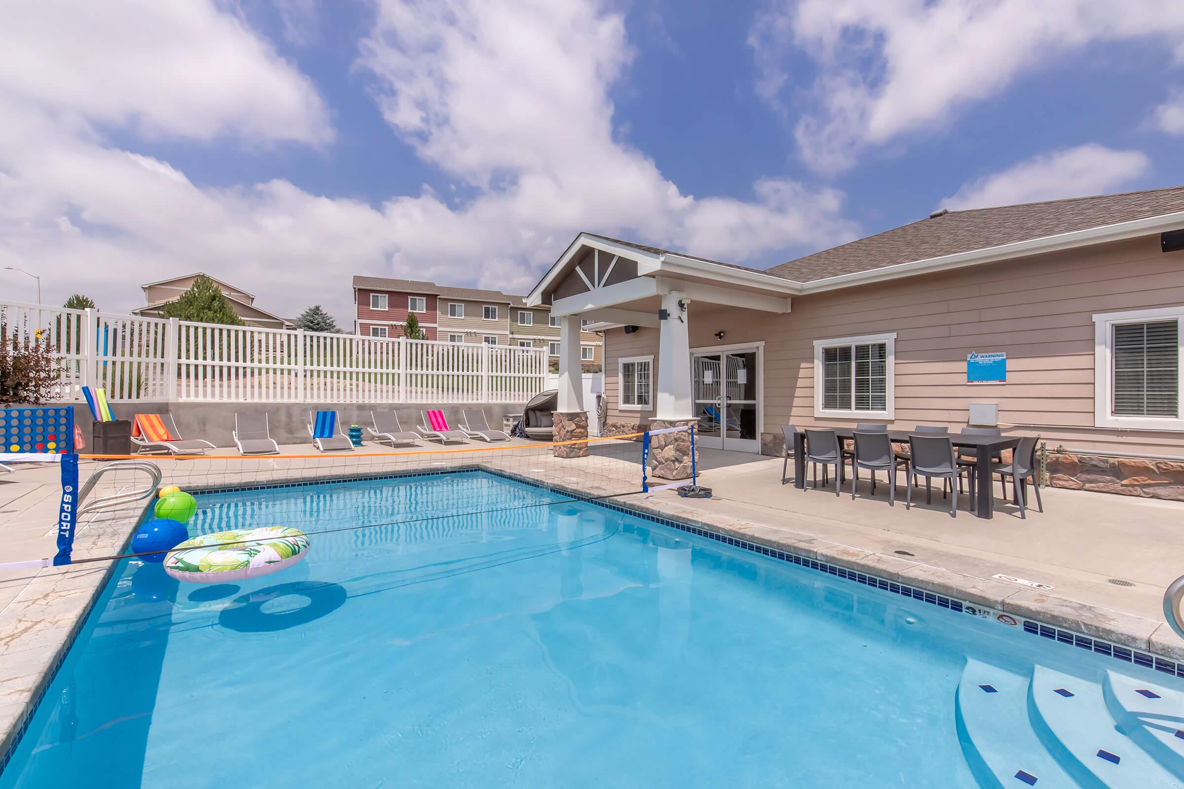 A clear blue swimming pool surrounded by lounge chairs, with a nearby shaded area. In the background, a building with multiple windows and a patio table set is visible. The sky is partly cloudy, creating a bright and inviting atmosphere.
