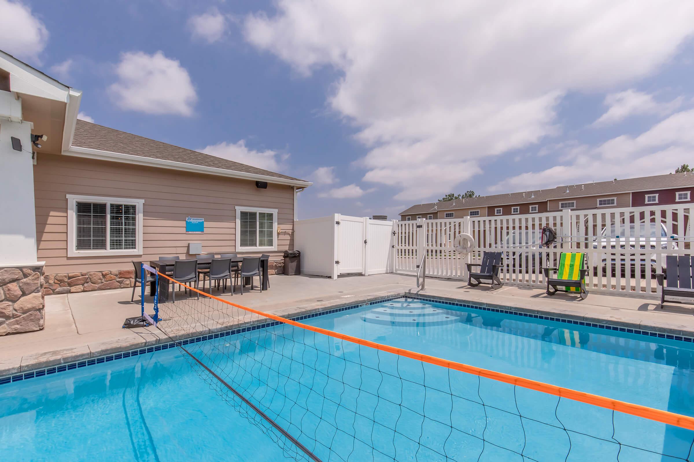 A sunny pool area featuring a clear blue swimming pool with a volleyball net, surrounded by lounge chairs. A beige building with windows is in the background, along with a fenced area. The sky is partly cloudy, creating a bright and inviting atmosphere.