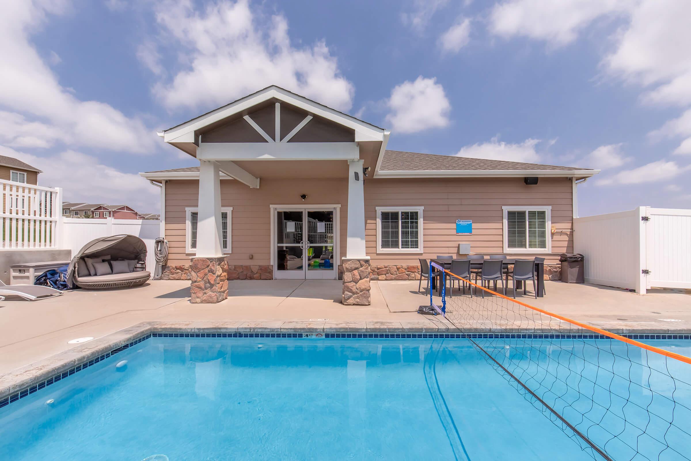 A residential backyard featuring a clear swimming pool with a volleyball net. In the background, there is a well-lit house with a porch and several windows. A cozy seating area is visible near the pool, along with a white fence surrounding the property. The sky is partly cloudy, creating a bright atmosphere.
