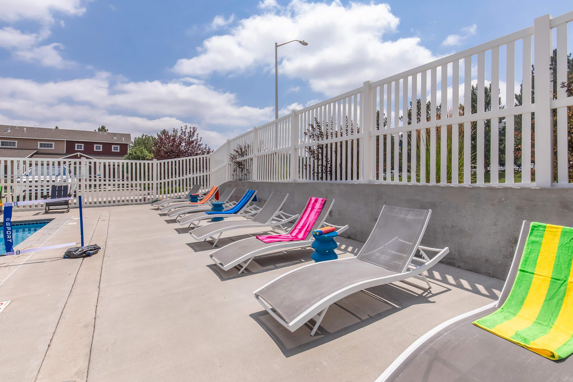 Lounge chairs in vibrant colors arranged by a swimming pool, with a white fence in the background and a clear blue sky dotted with clouds. A few towels are draped over the chairs, creating a relaxed, inviting atmosphere for sunbathing and leisure.