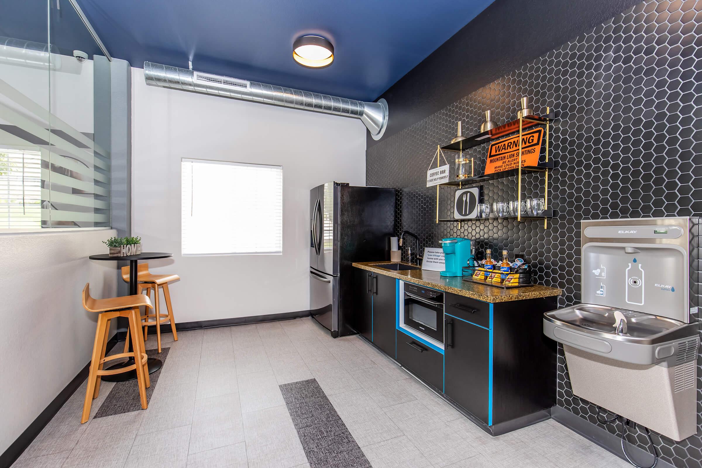 Modern kitchen area featuring a sleek black and gold counter with a microwave, refrigerator, and snacks. There are two wooden barstools, a water fountain, and a window providing natural light. The wall has a stylish hexagonal pattern and there are decorative items displayed on shelves.
